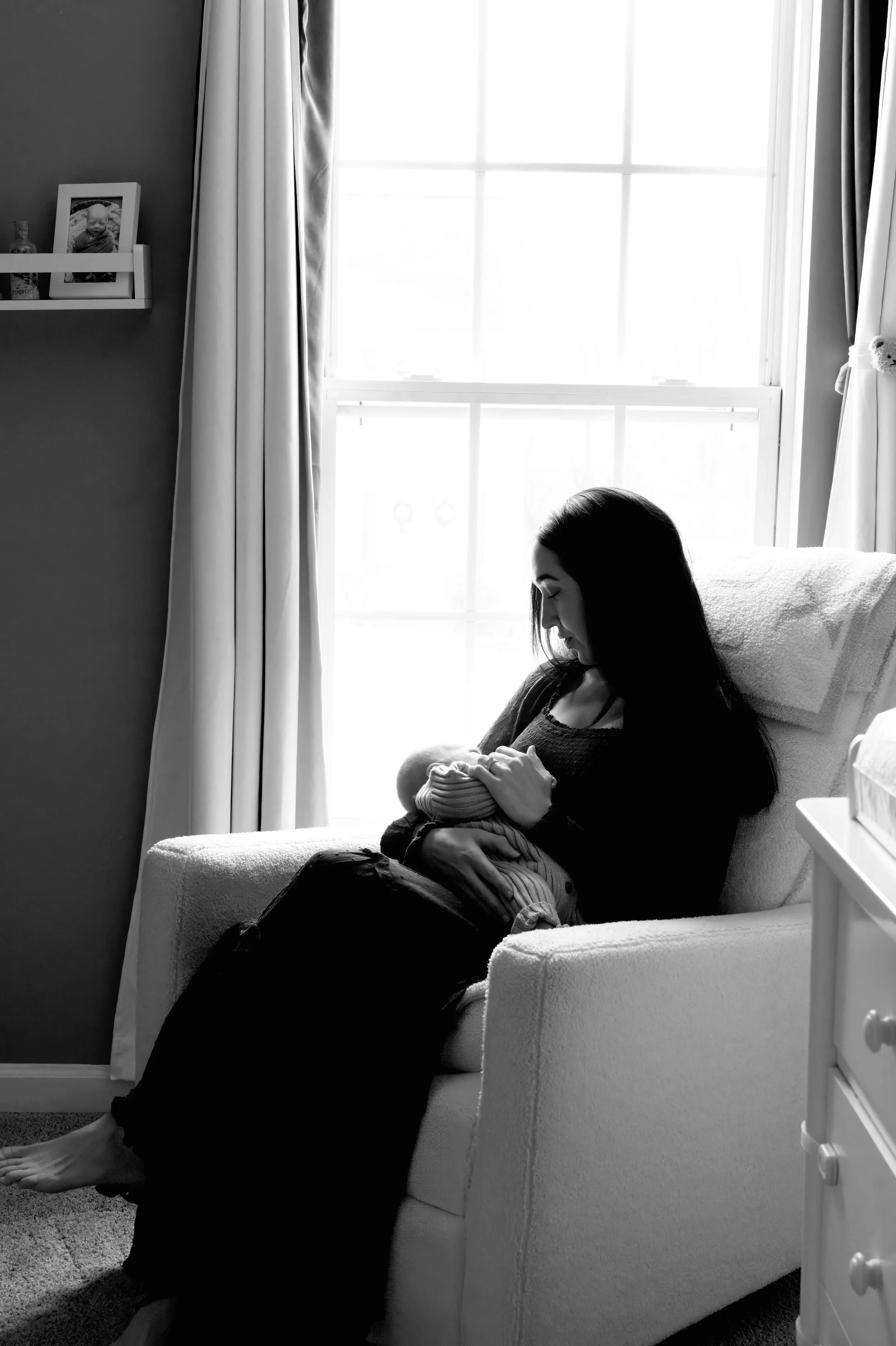 A woman with long dark hair sitting on a white armchair during the daytime, holding and breastfeeding a baby, in a room with a window, curtains, a shelf with framed photos, and some decorative items.