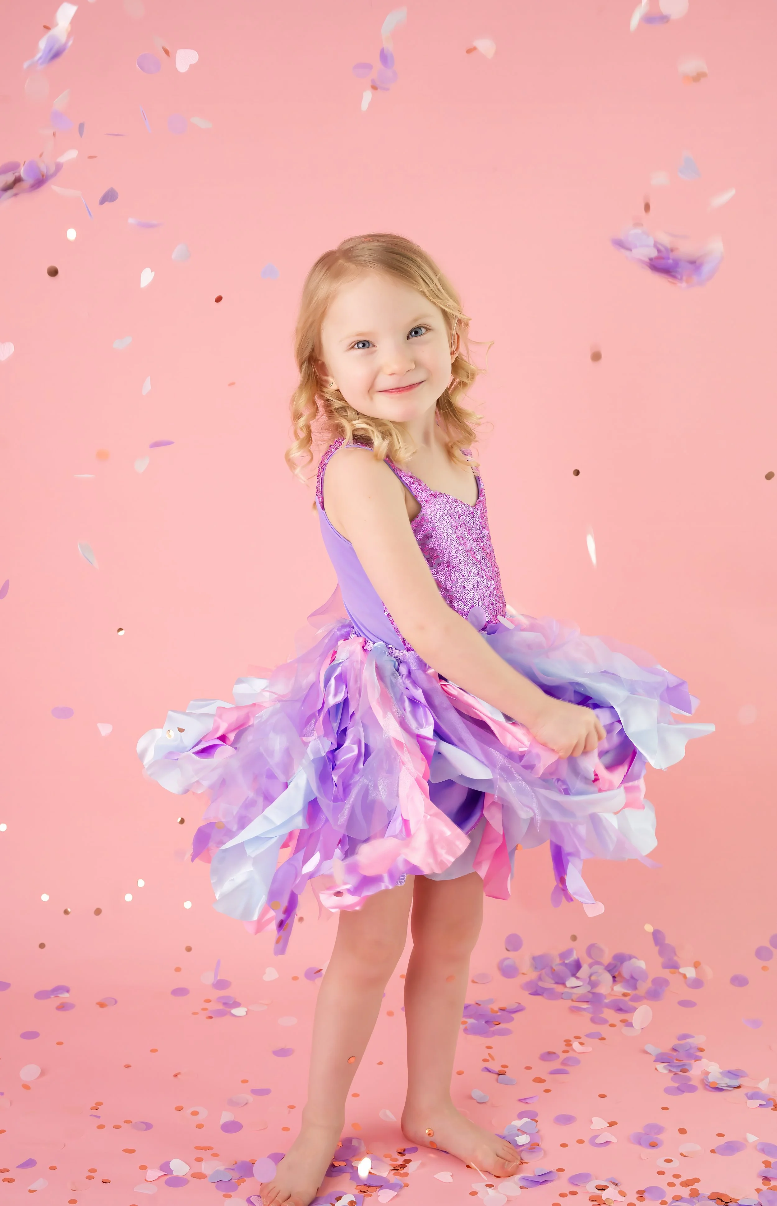 A young girl with blonde curly hair wearing a purple and pink tutu dress, standing barefoot on a pink background with confetti falling around her.