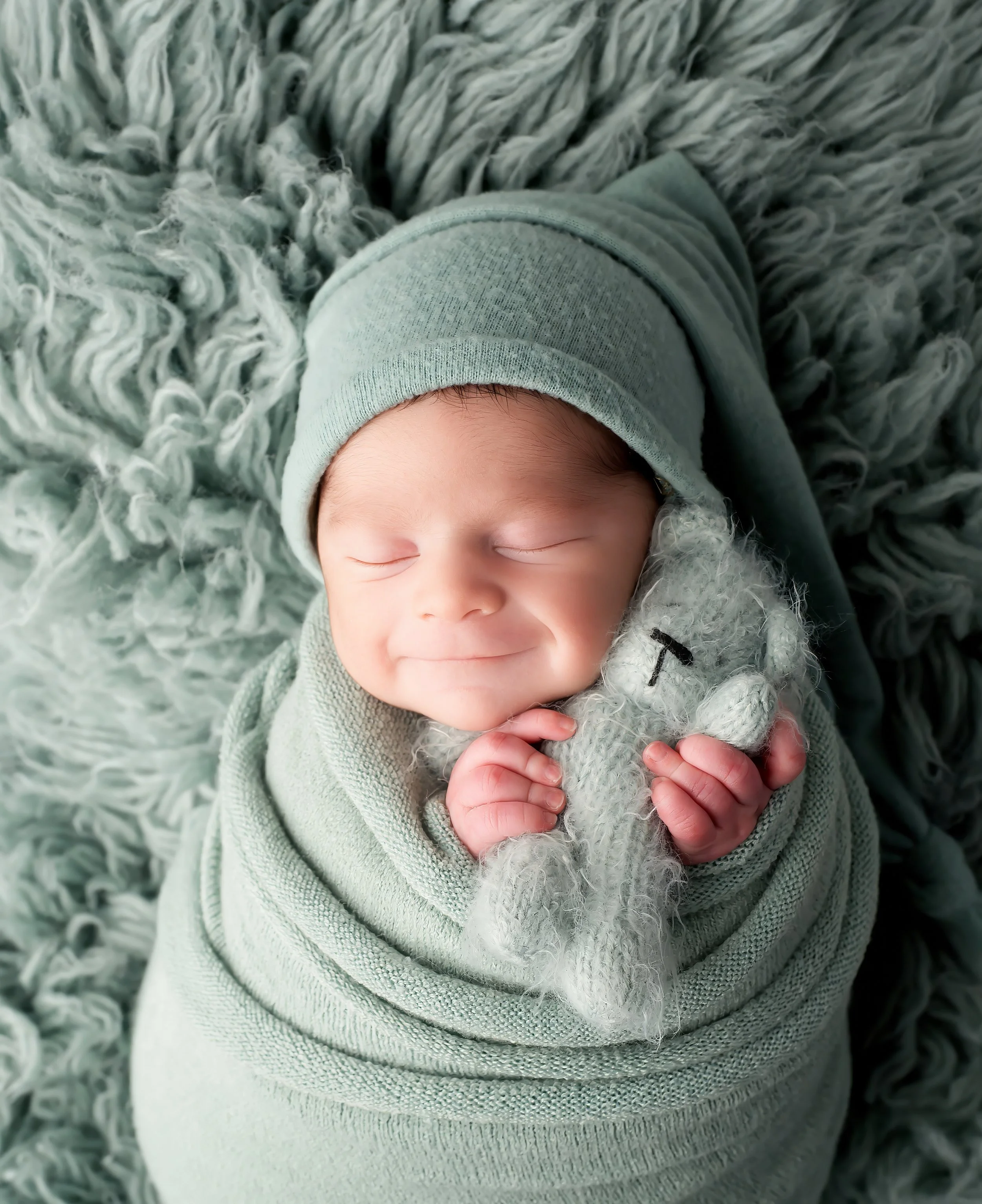 Close-up of a smiling baby with closed eyes, wearing a light dusty sage knit hat and surrounded by a soft, curly dusty sage flokati rug.