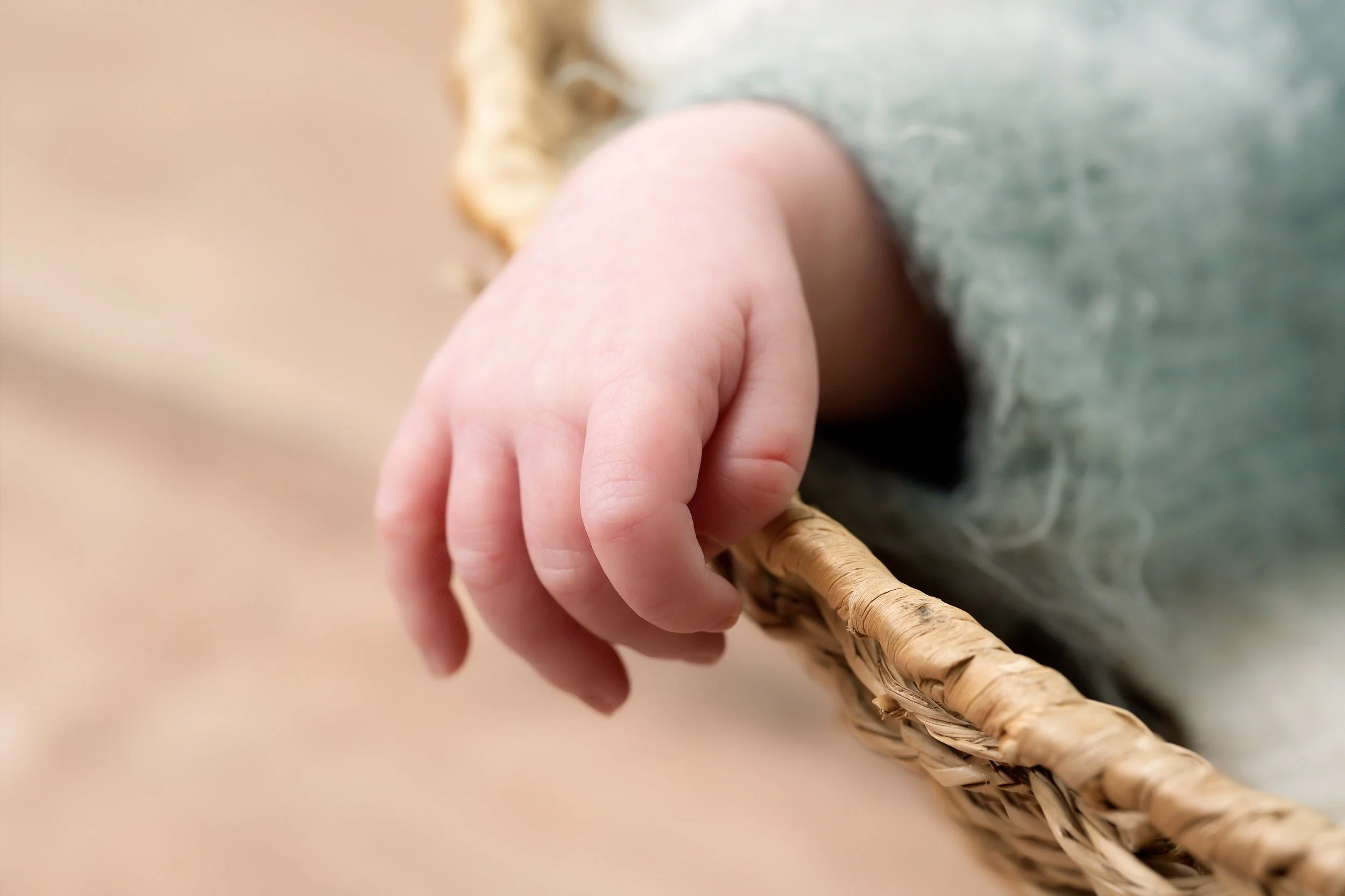 A close-up of a baby's hand holding a woven wicker basket handle with a dusty sage piece of fabric and a beige surface background.