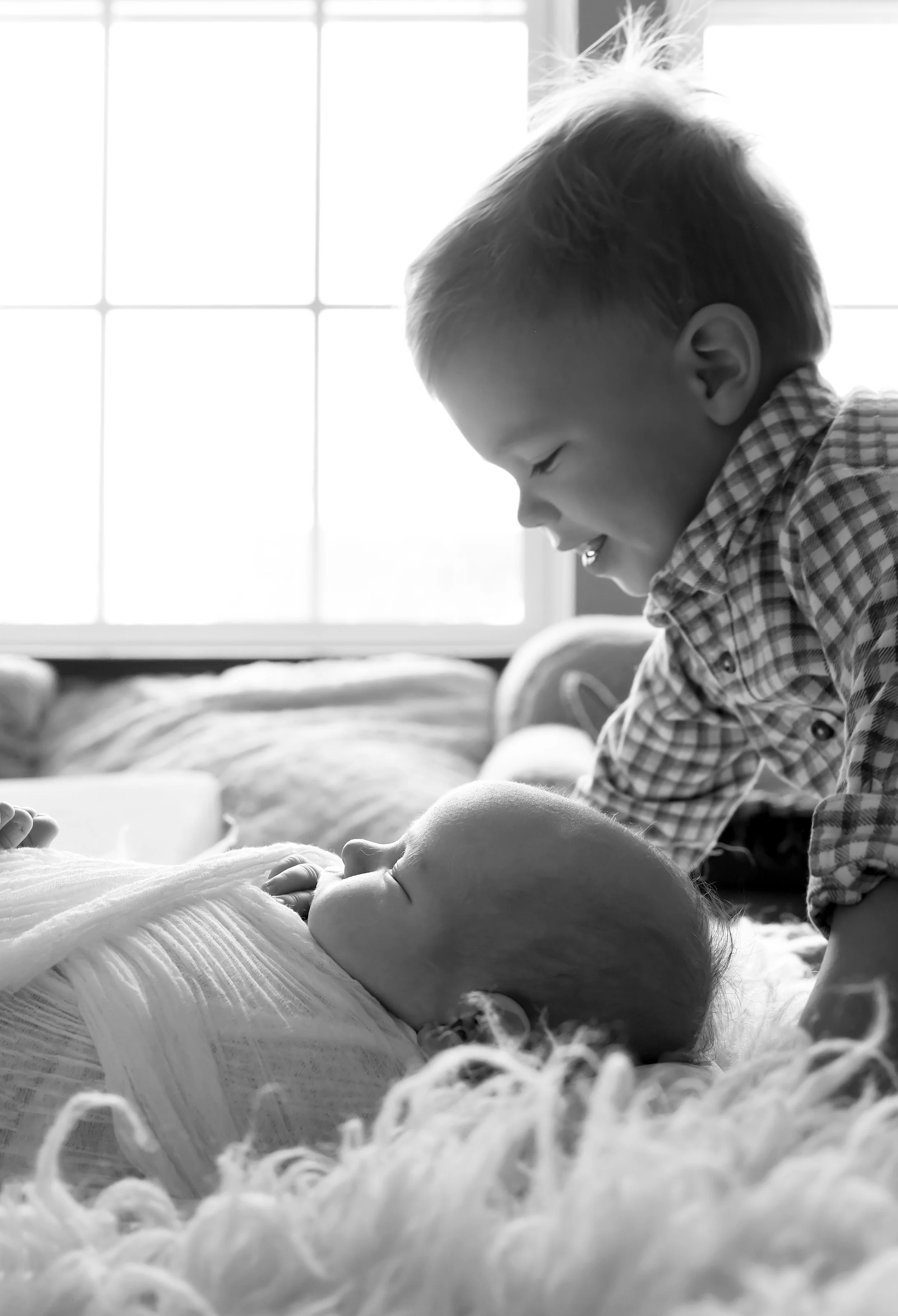 A young boy with short hair smiling and leaning over a sleeping baby who is lying on a bed or blanket. The background shows a window with sunlight coming through.