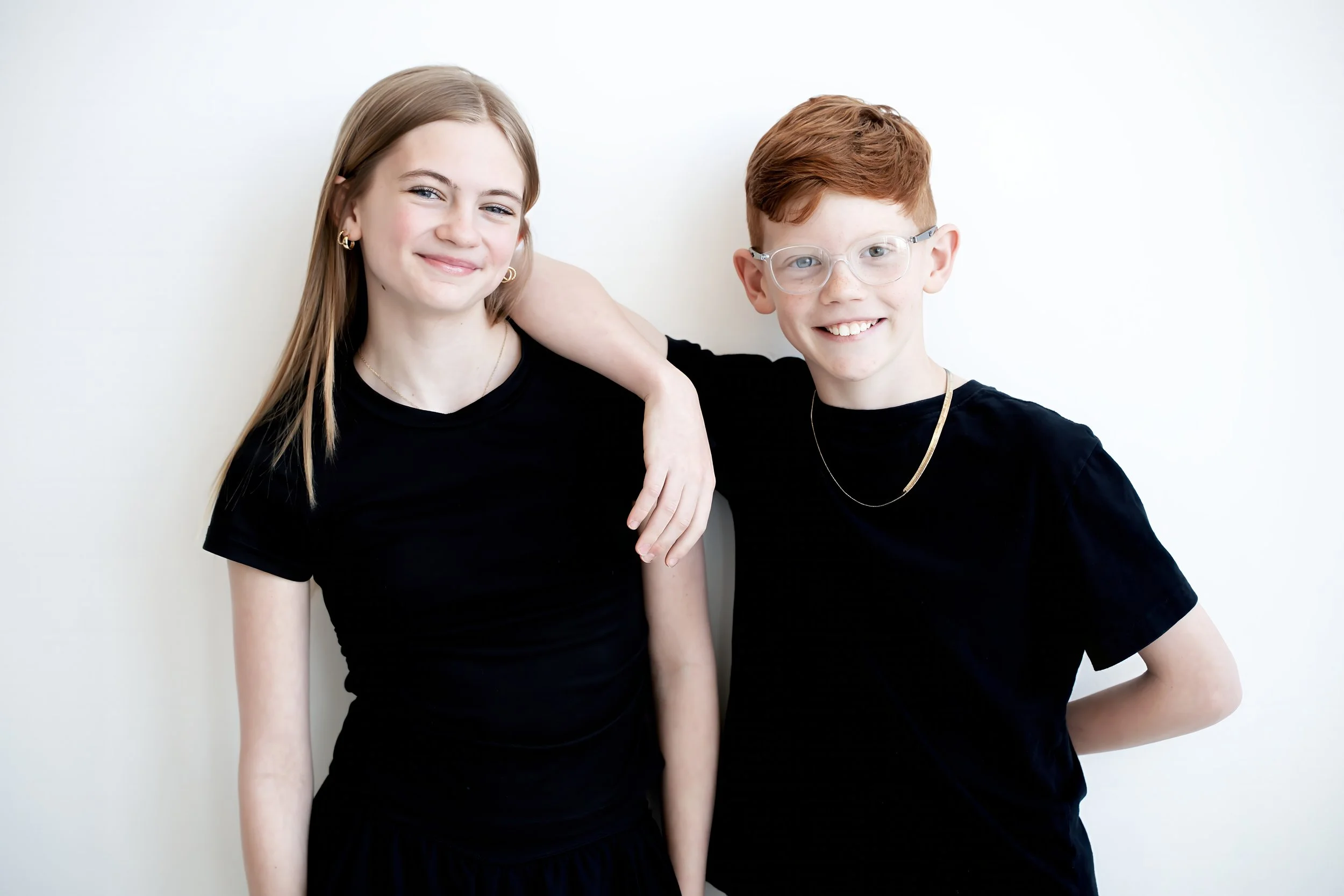 Two young people, a girl with long blonde hair and a boy with short red hair and glasses, standing close together and smiling, against a plain white background