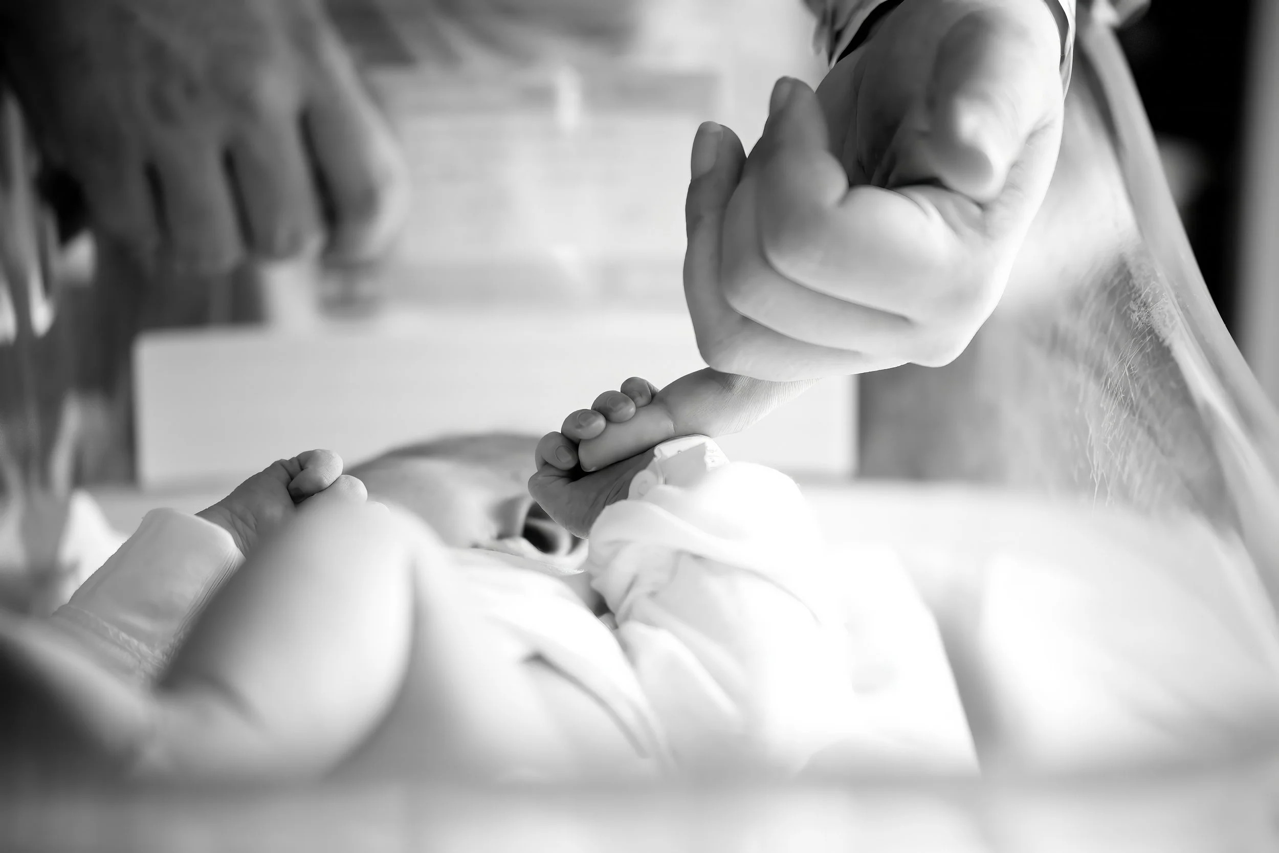 Close-up of a adult's pinky being held by a newborn with a protective grip. The background shows other hands, hinting at a group activity or support scene, in black and white.