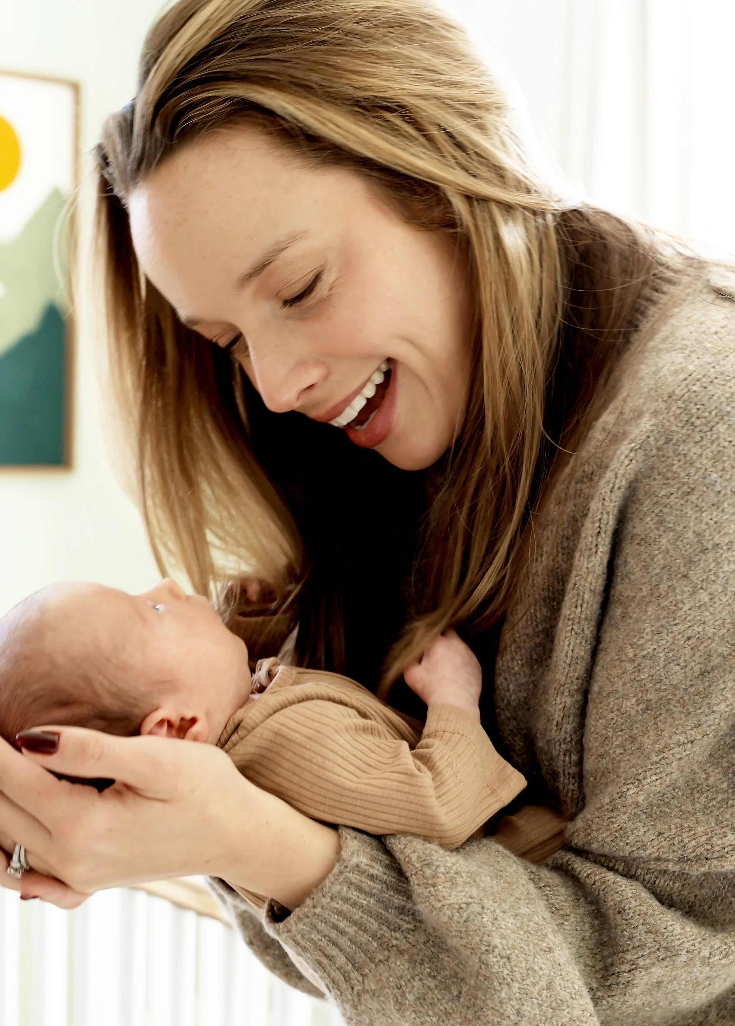 A woman with long, light brown hair smiling and looking down at a newborn baby she is holding in her arms. The woman is wearing a beige sweater and the baby is dressed in a beige striped outfit.