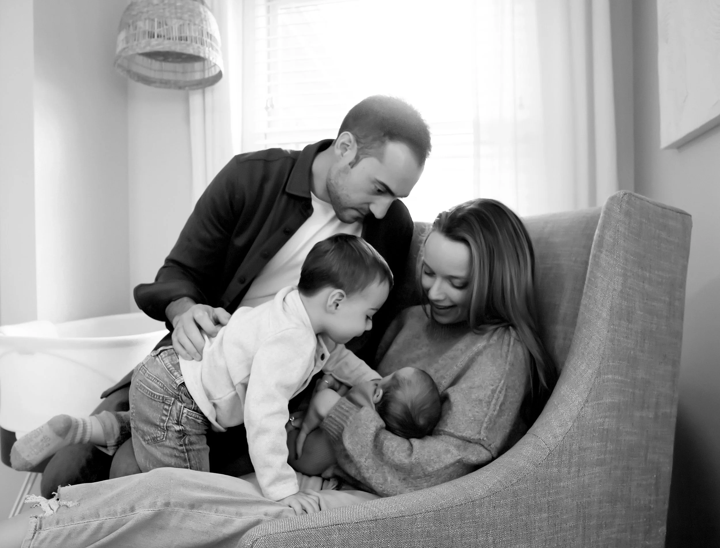 Black and white photo of a family of five gathered around and cuddling on a couch. The father and mother are leaning over, engaging with their two young children.