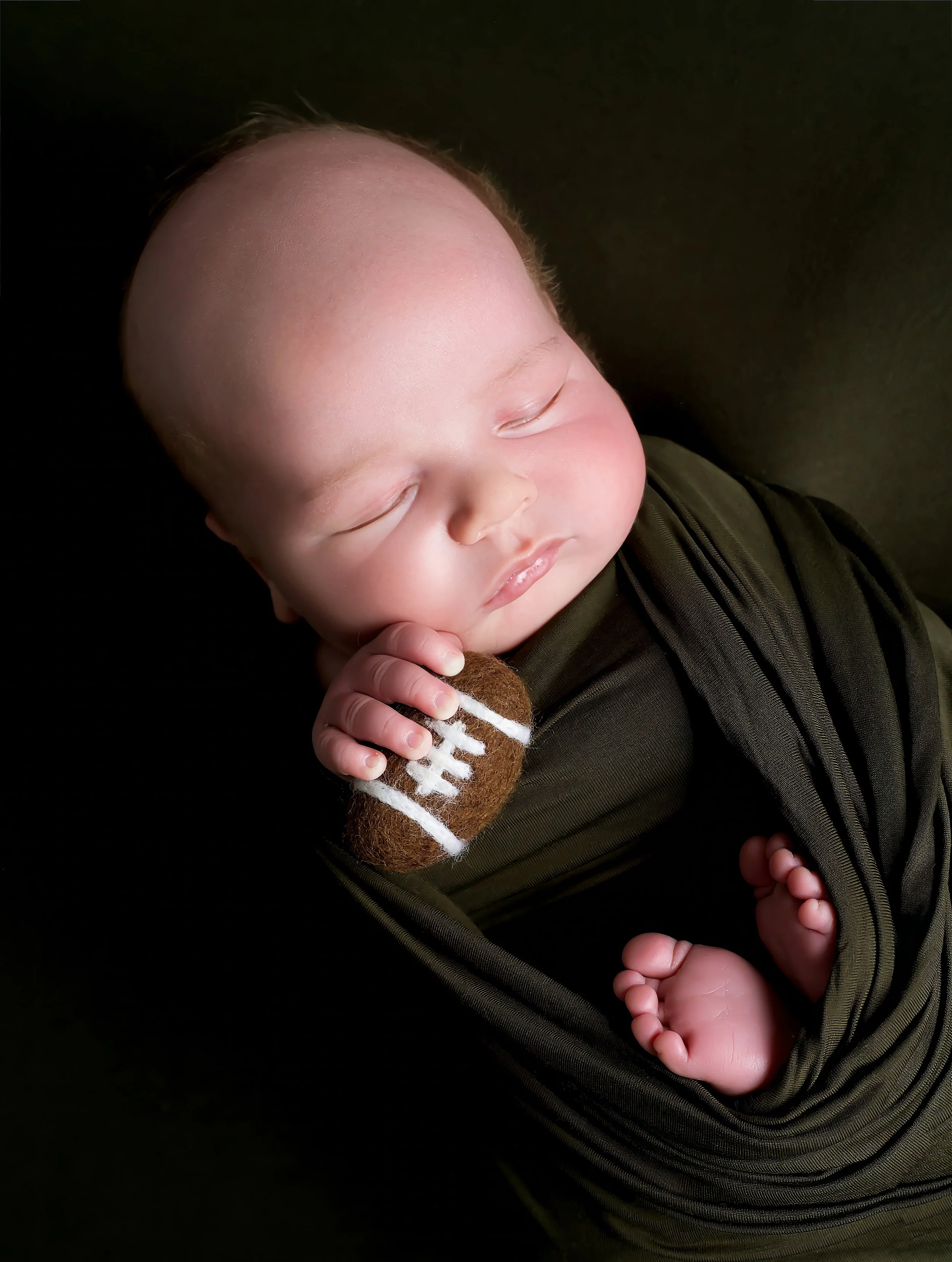 A sleeping baby with closed eyes, a bald head, and pink cheeks, holding a small stuffed animal in a dark green stroller or carrier against a black background.