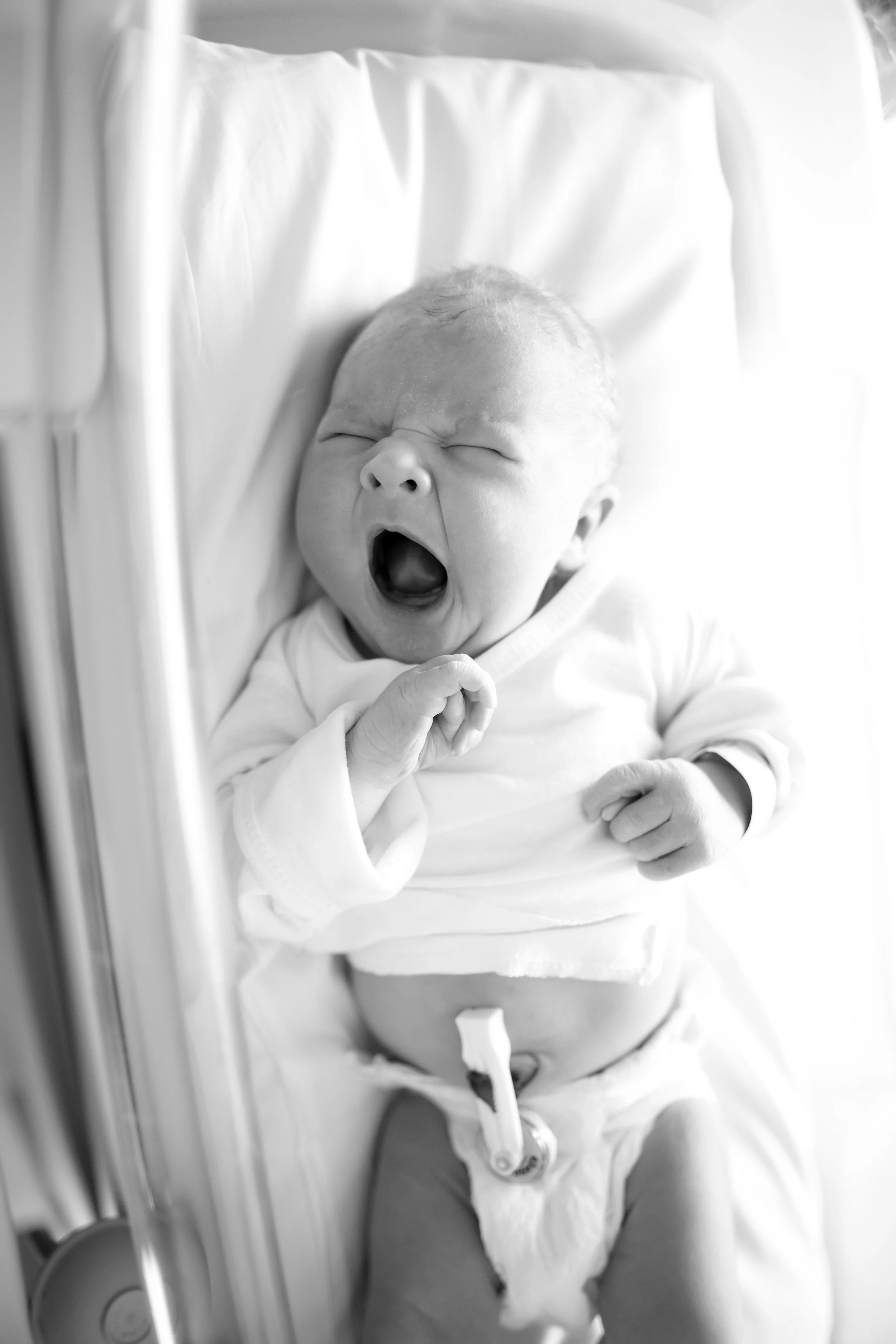 Black and white photo of a newborn baby yawning while lying in a hospital bassinet, dressed in a white outfit with a pacifier clip attached.