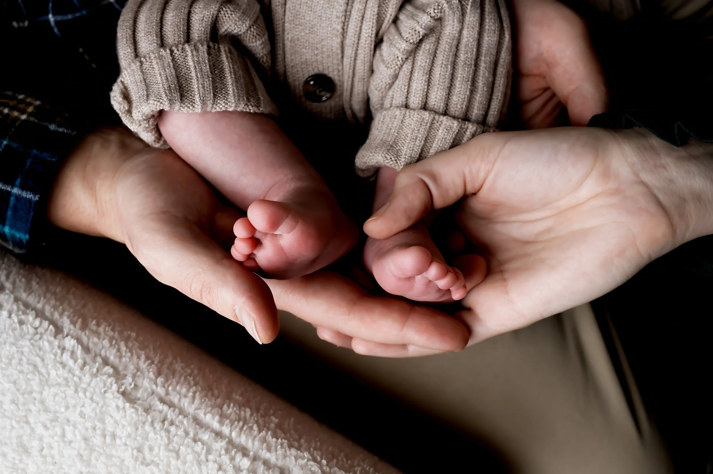 Close-up of adult and baby hands holding tiny baby feet, with a beige sweater sleeve visible.
