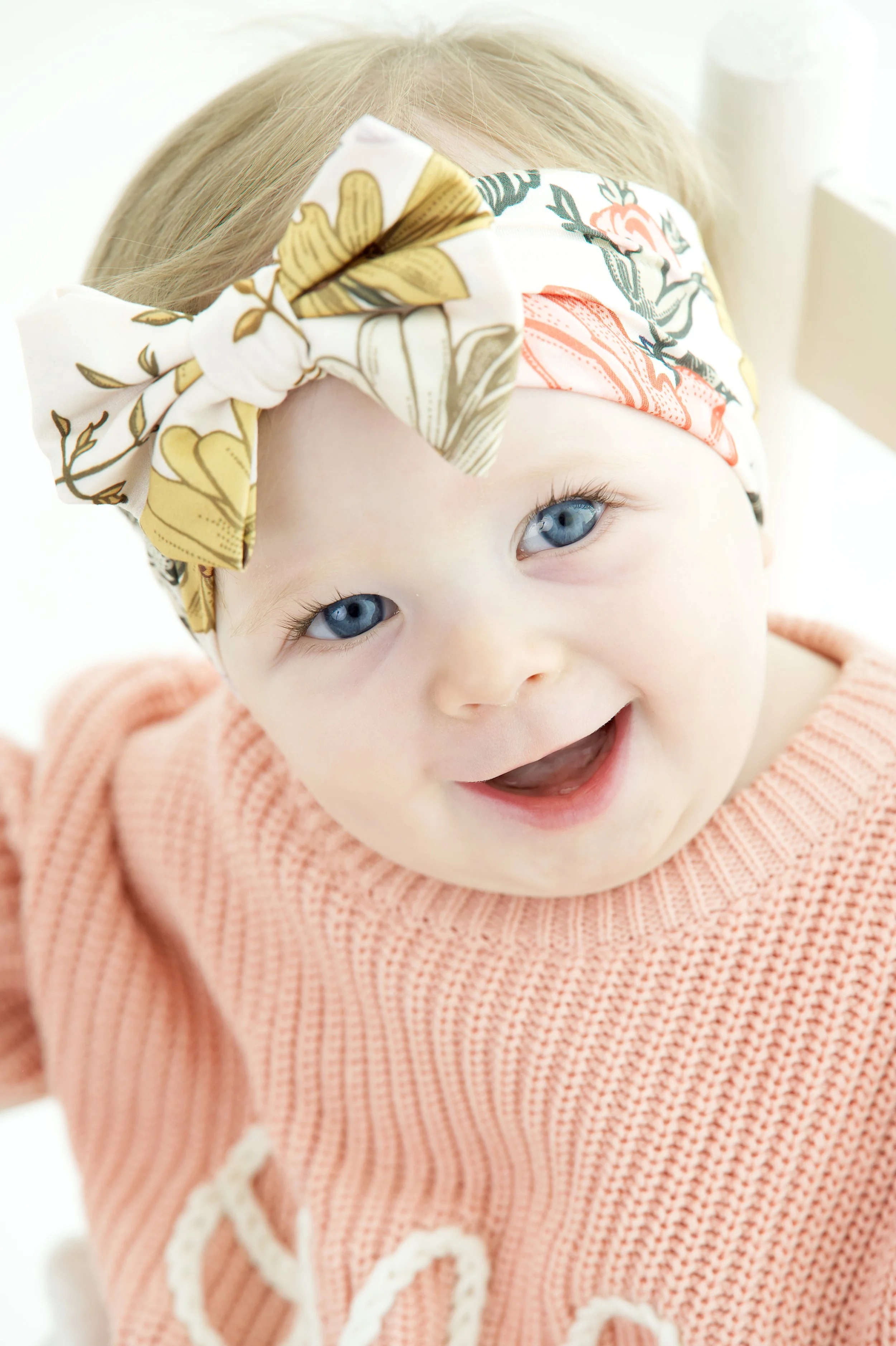 A smiling baby with blue eyes wearing a peach knitted sweater and a colorful patterned headband.