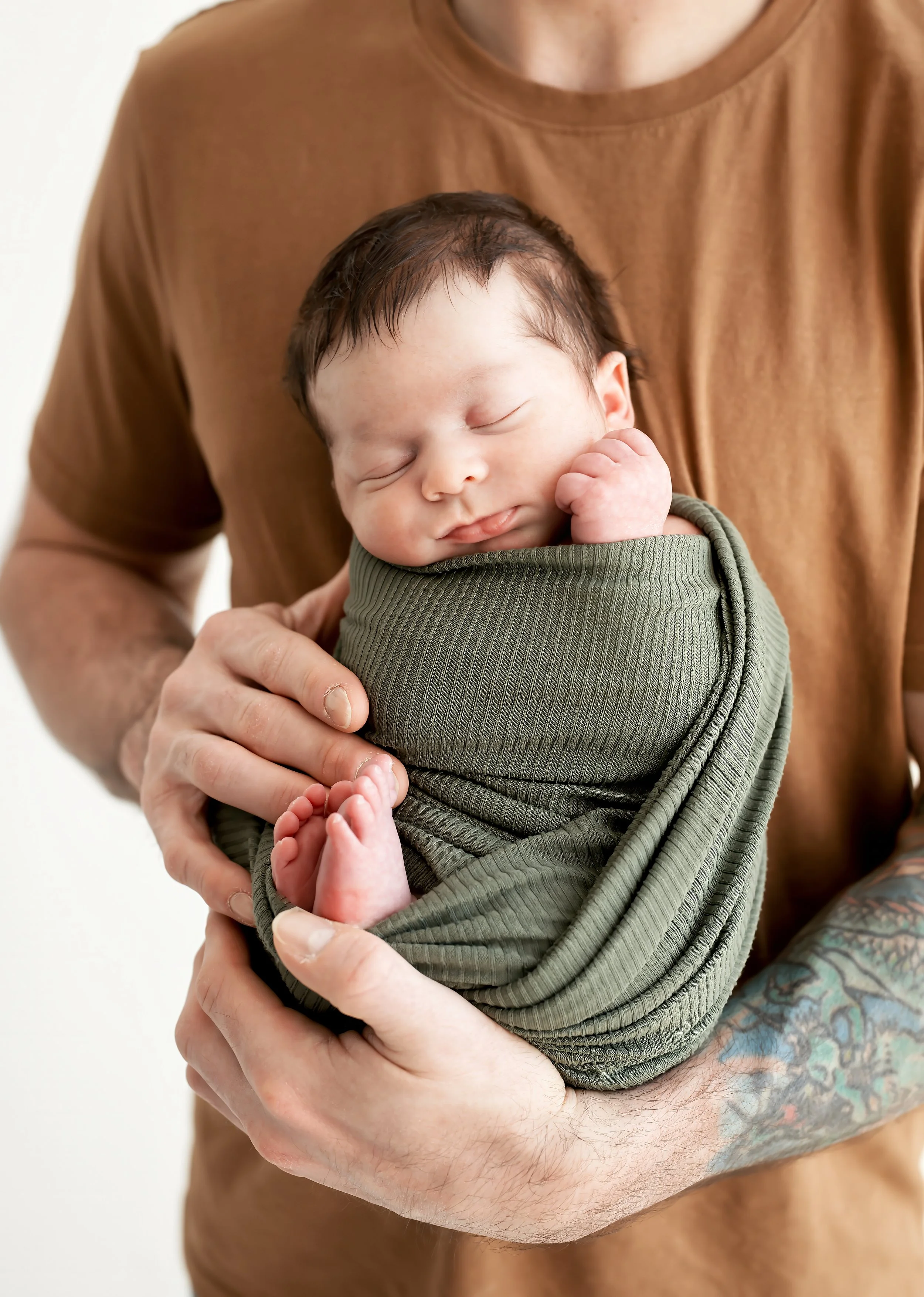 Close-up of a person holding a sleeping baby wrapped in green fabric, with the person's arms and hands supporting the baby, and a tattoo visible on one arm.