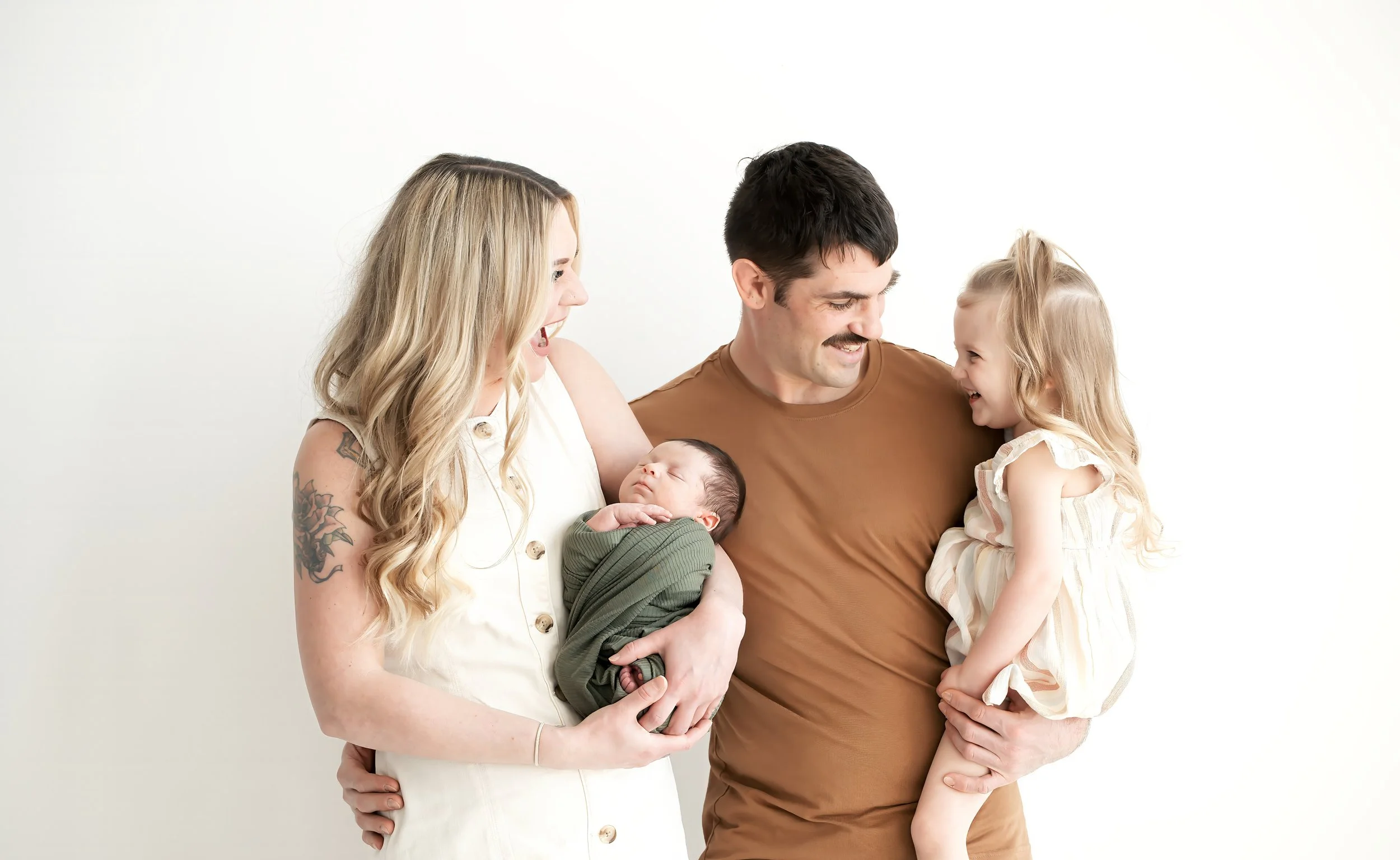 A family of four, including a woman with long blonde hair and tattoos, a man with short dark hair and a mustache, a young girl with blonde hair, and a newborn baby, are happy and smiling together against a white background.