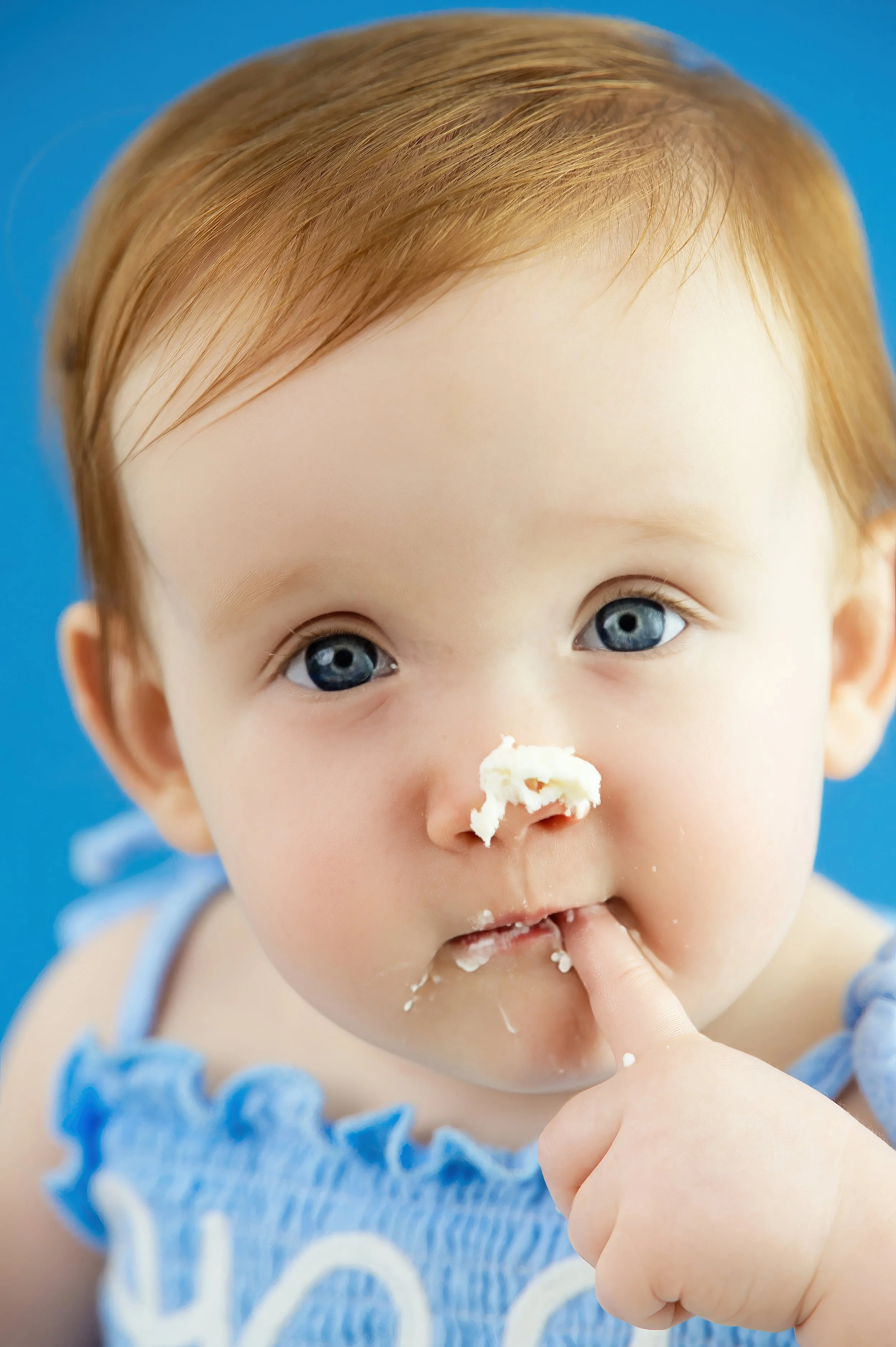 Close-up of a young child with blue eyes and light brown hair, eating cake with white frosting. The child has frosting on their nose and finger, and is wearing a light blue outfit.