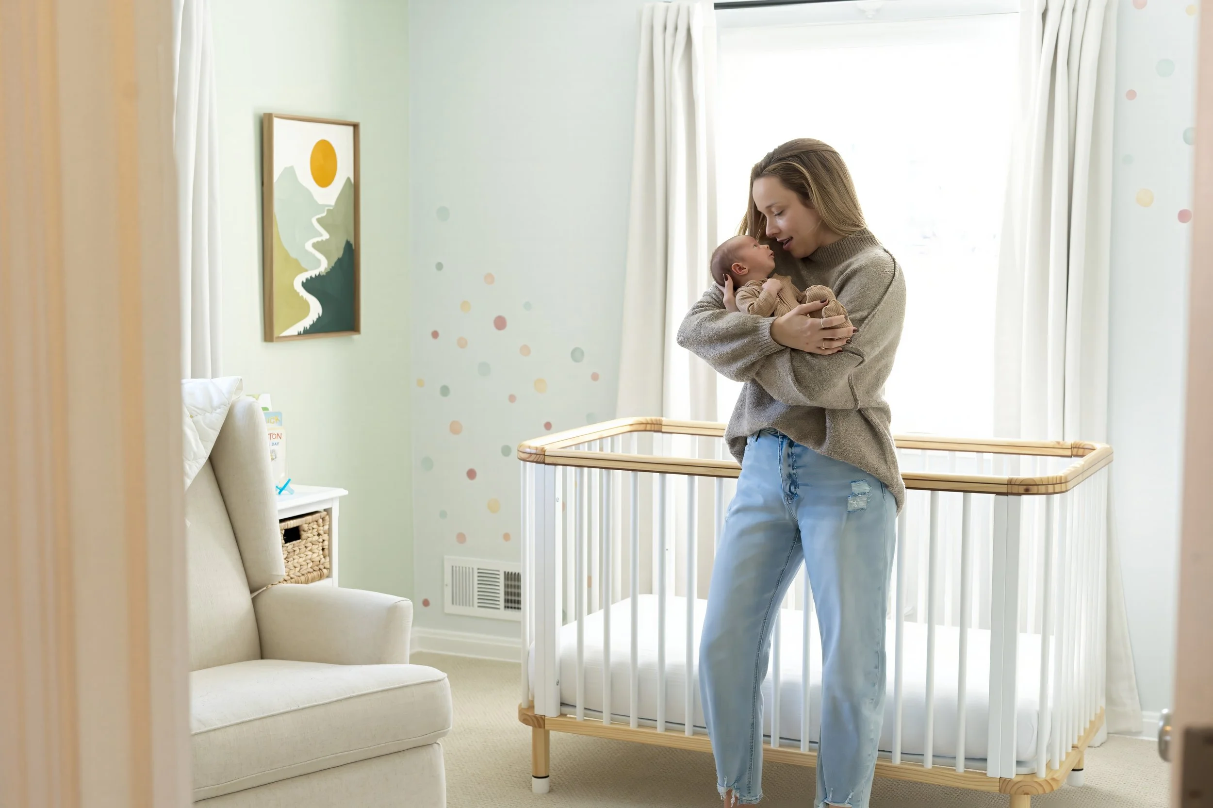 A woman holding a newborn baby in a nursery with a crib, a beige armchair, and wall art featuring a mountain landscape and a sun.