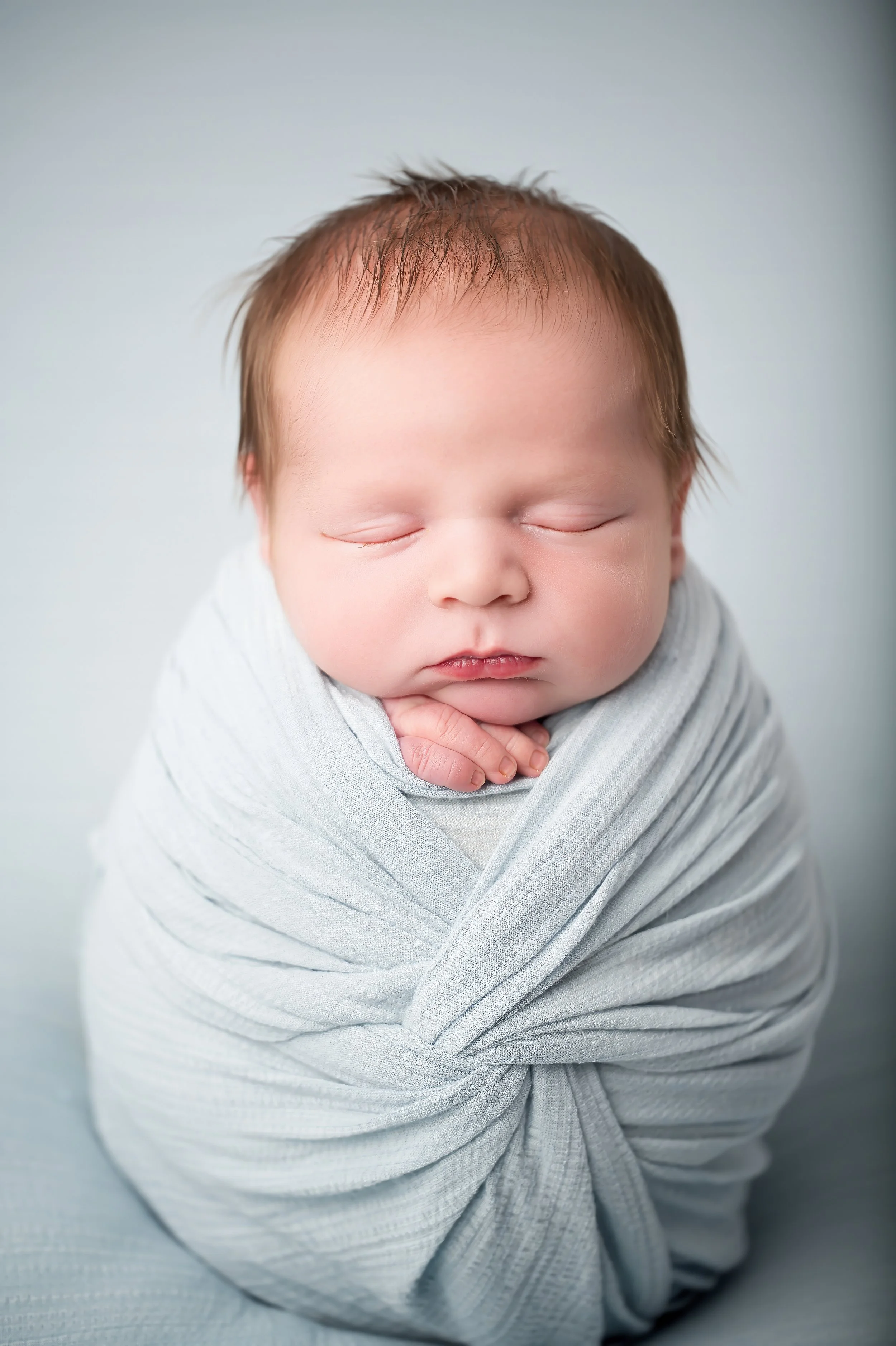 Close-up of a newborn baby with closed eyes, wrapped in a light gray blanket, with soft lighting and a plain background.