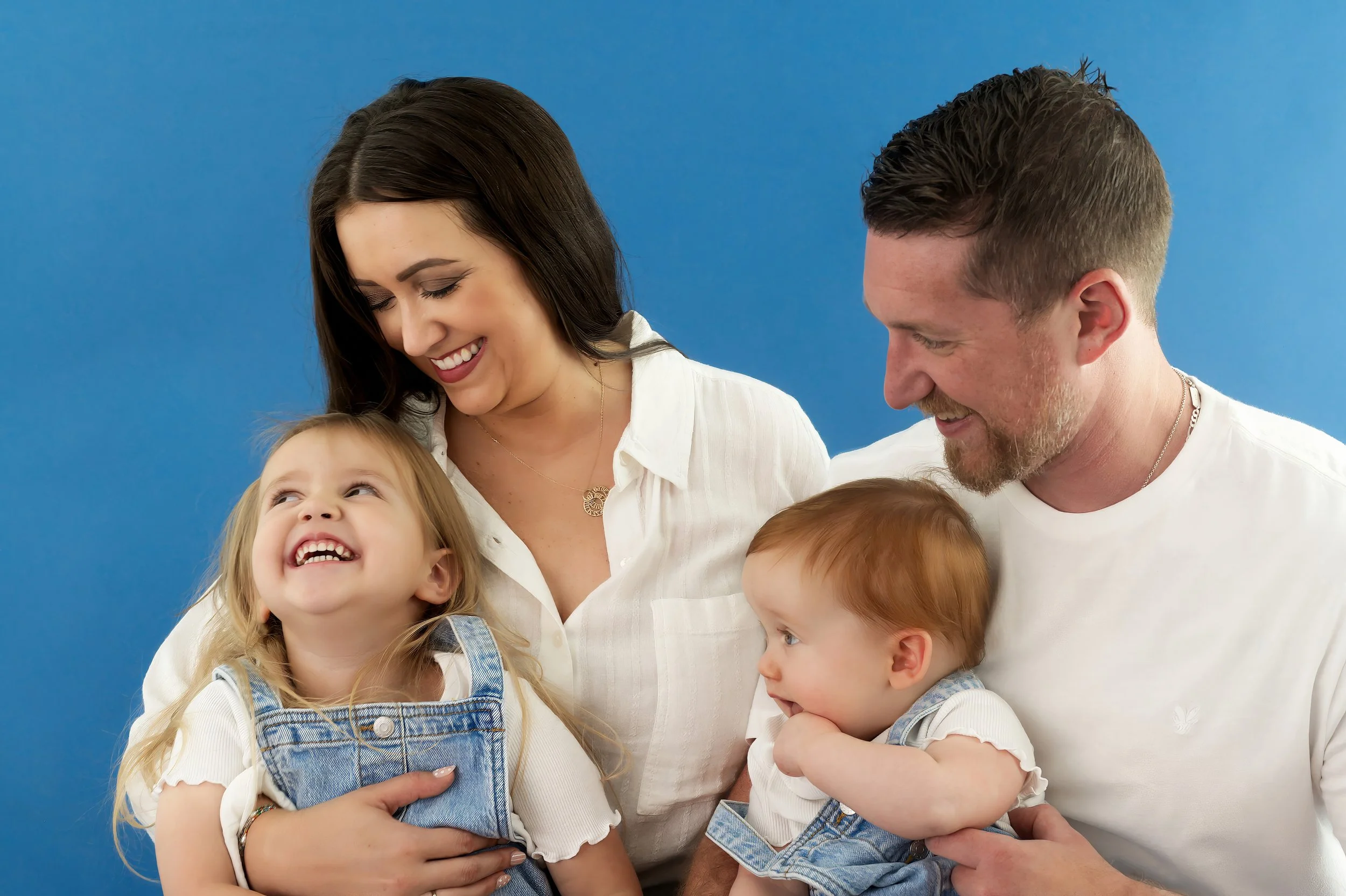 A husband and wife with two young children smiling and looking at each other against a solid blue background.