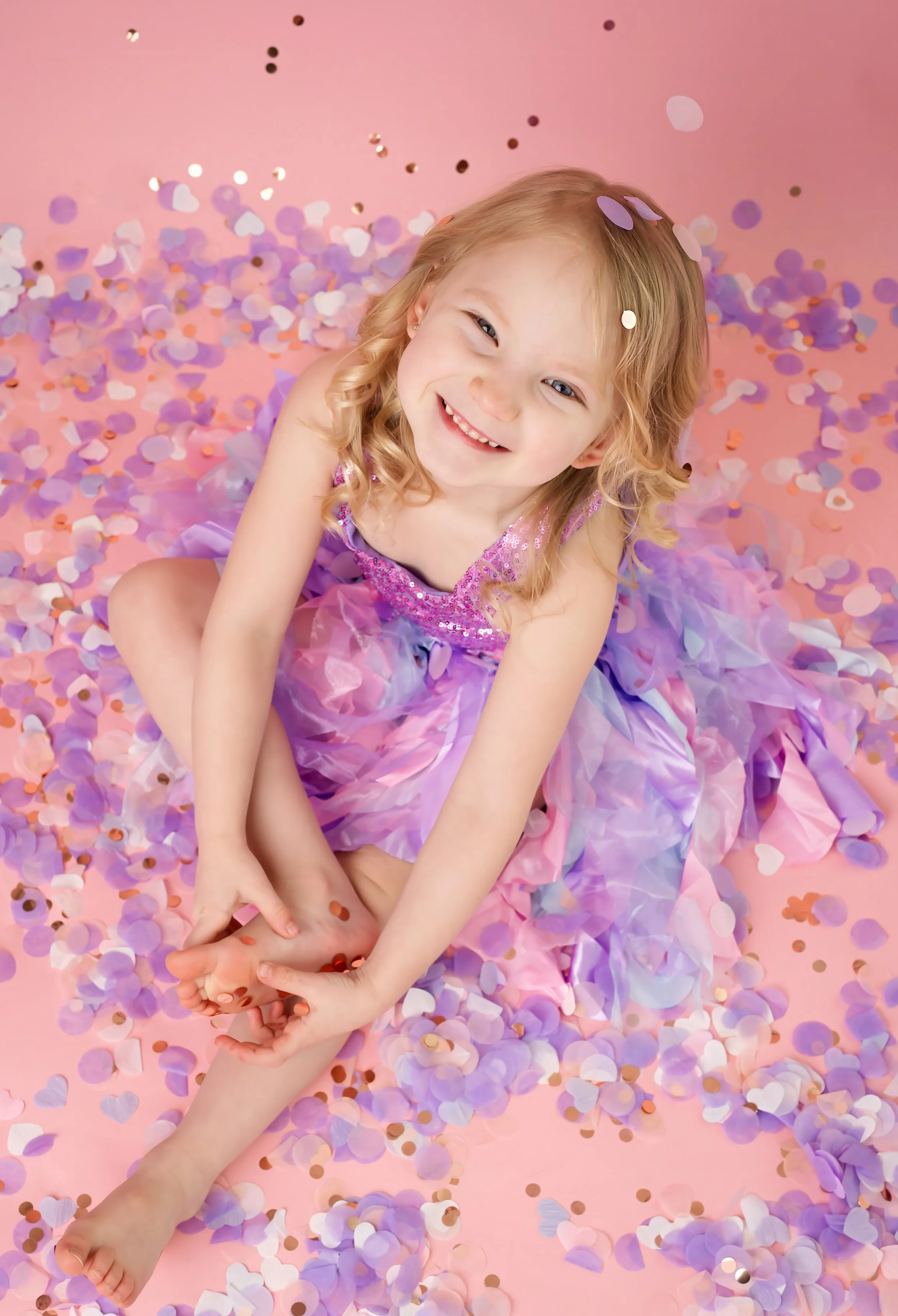 Smiling young girl with blonde hair in a pink and purple dress sitting on a pink background, surrounded by purple, white, pink, and gold confetti shaped like hearts and circles.