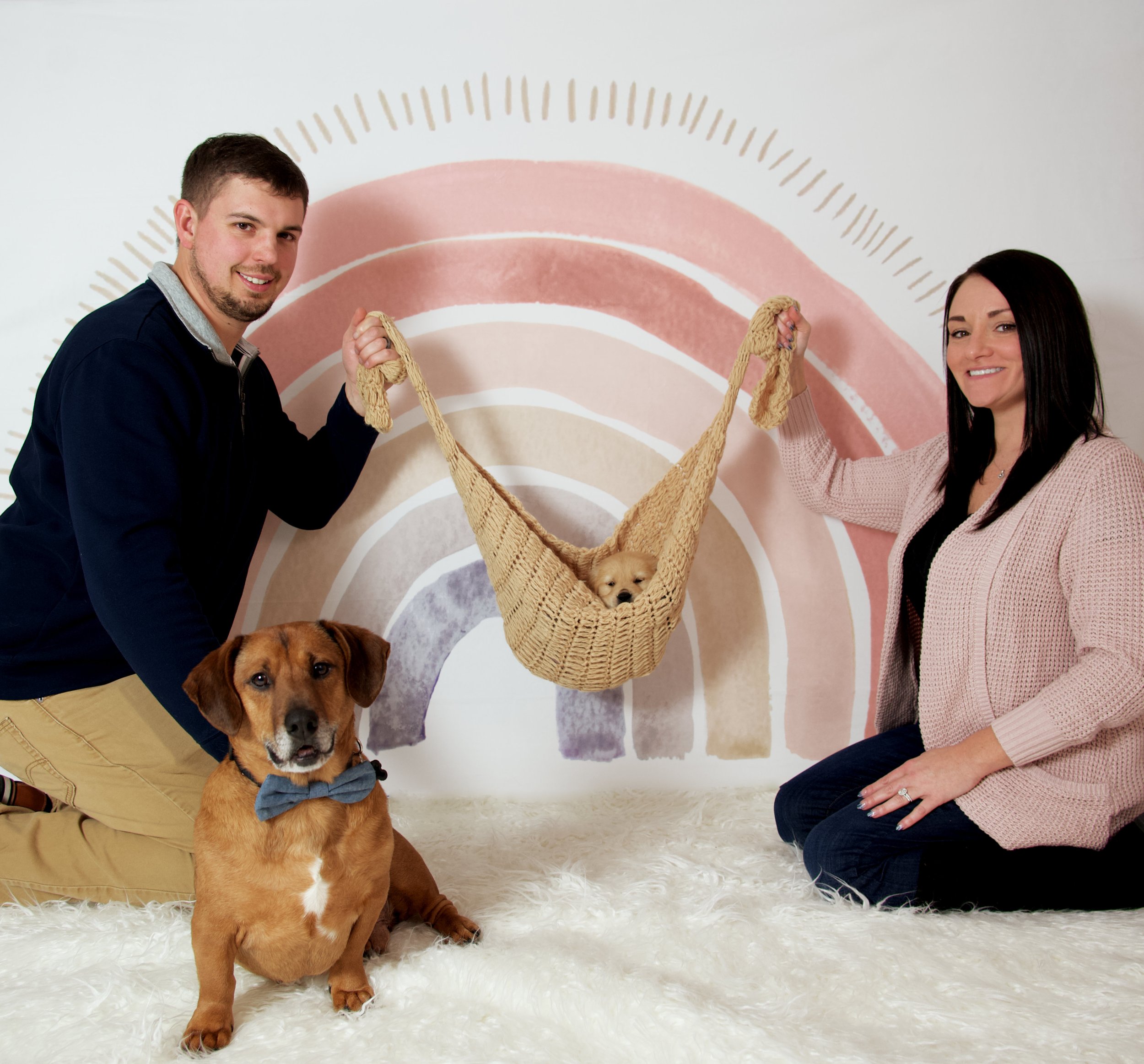 A man and woman are kneeling on a furry rug in front of a pastel rainbow wall. They are holding a small woven basket hammock containing a puppy. There is also a brown dog wearing a bow tie sitting on the rug.