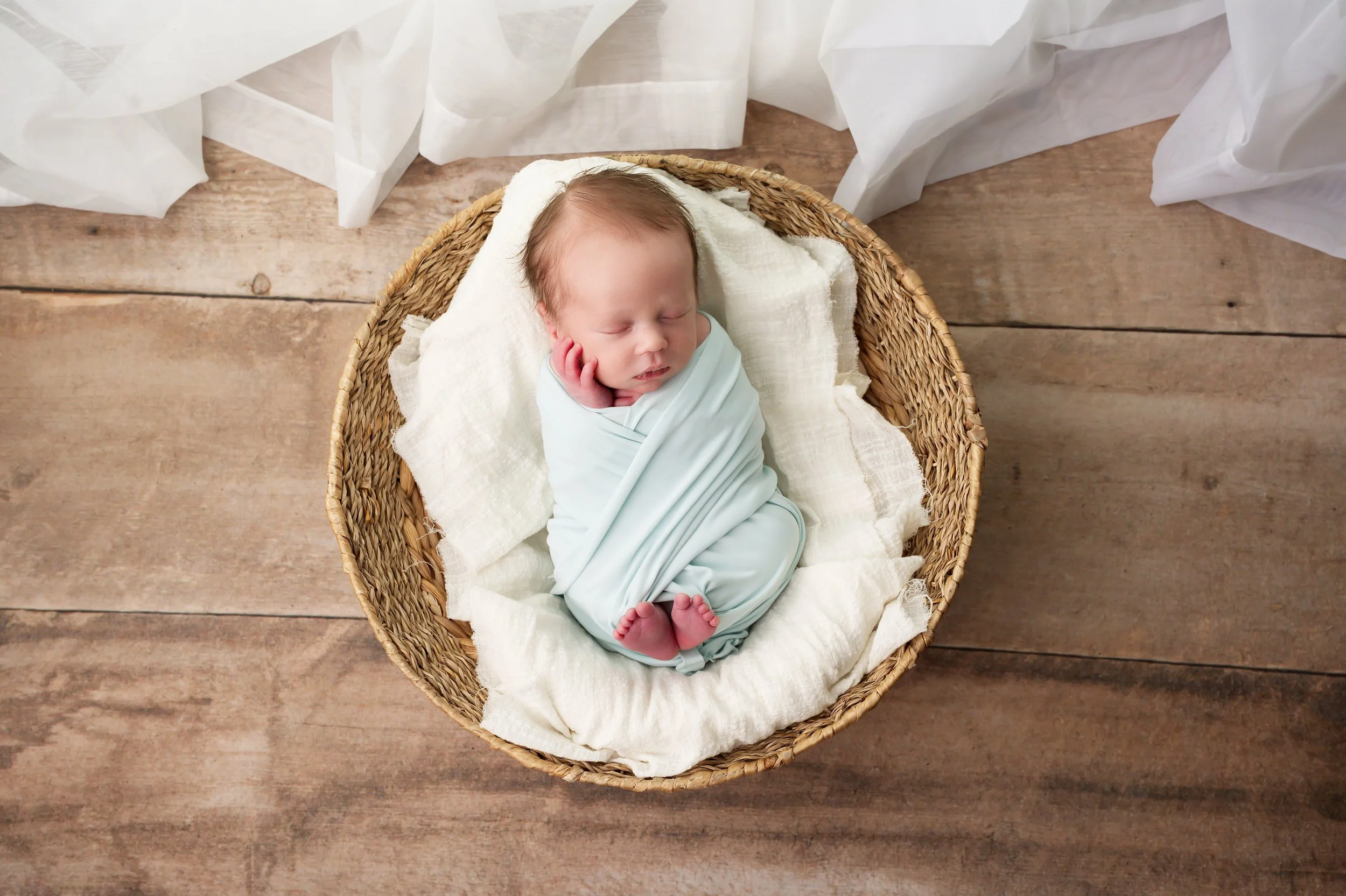 Smiling newborn baby resting in a wicker basket, wrapped in a light blue swaddle, on a wooden floor with sheer white fabric around.