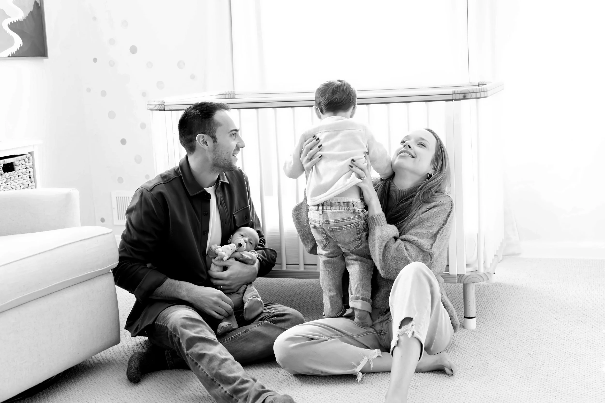A family of four, including a man, woman, a young boy, and a baby, gathered in a bright room with a crib. The woman is holding the young boy, who is facing away, while the man is sitting on the floor holding the baby. The woman and man are smiling at