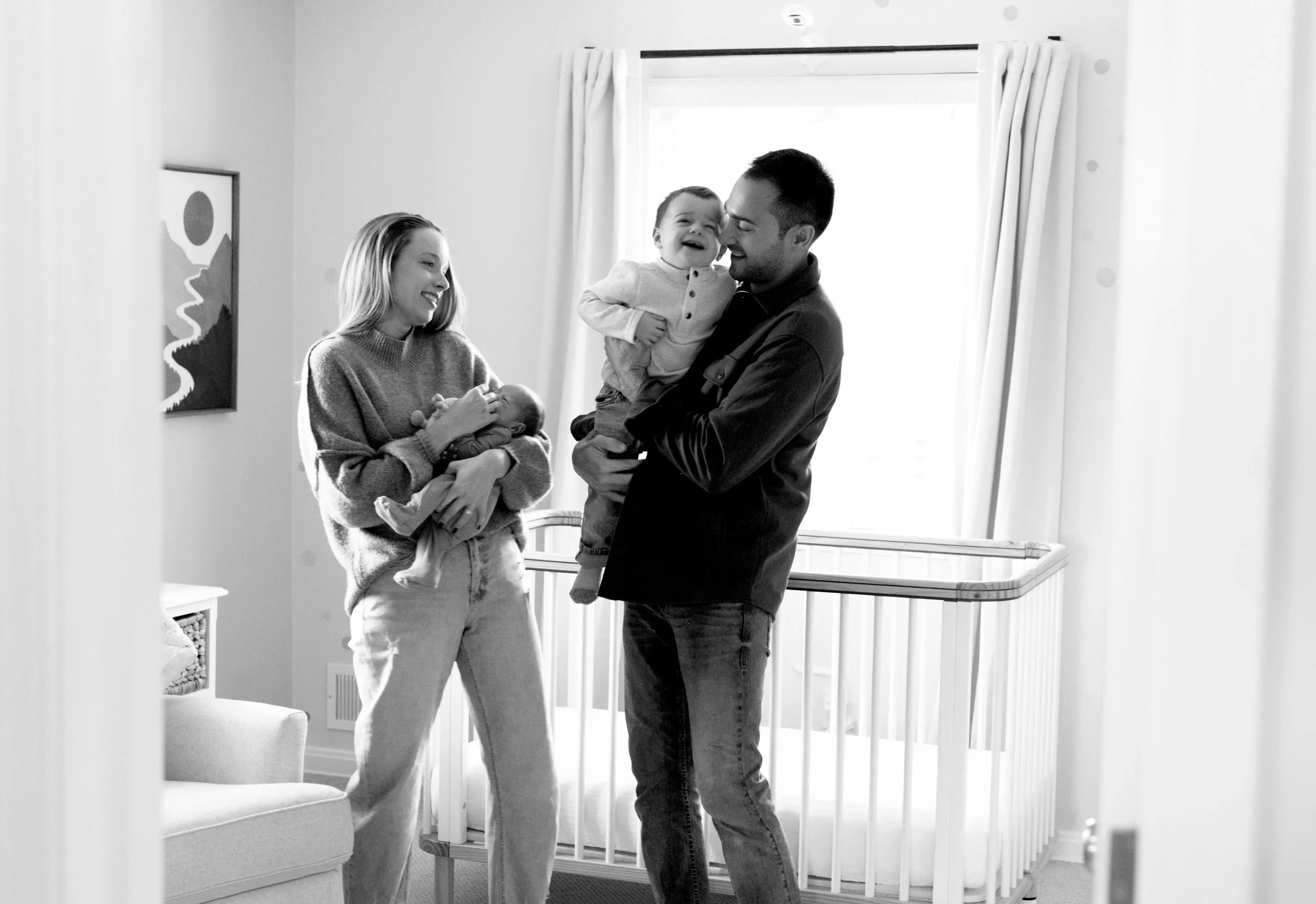 A black and white photo of a happy family with two women and two children, one being carried by each woman, in a well-lit living room with a window and curtains.