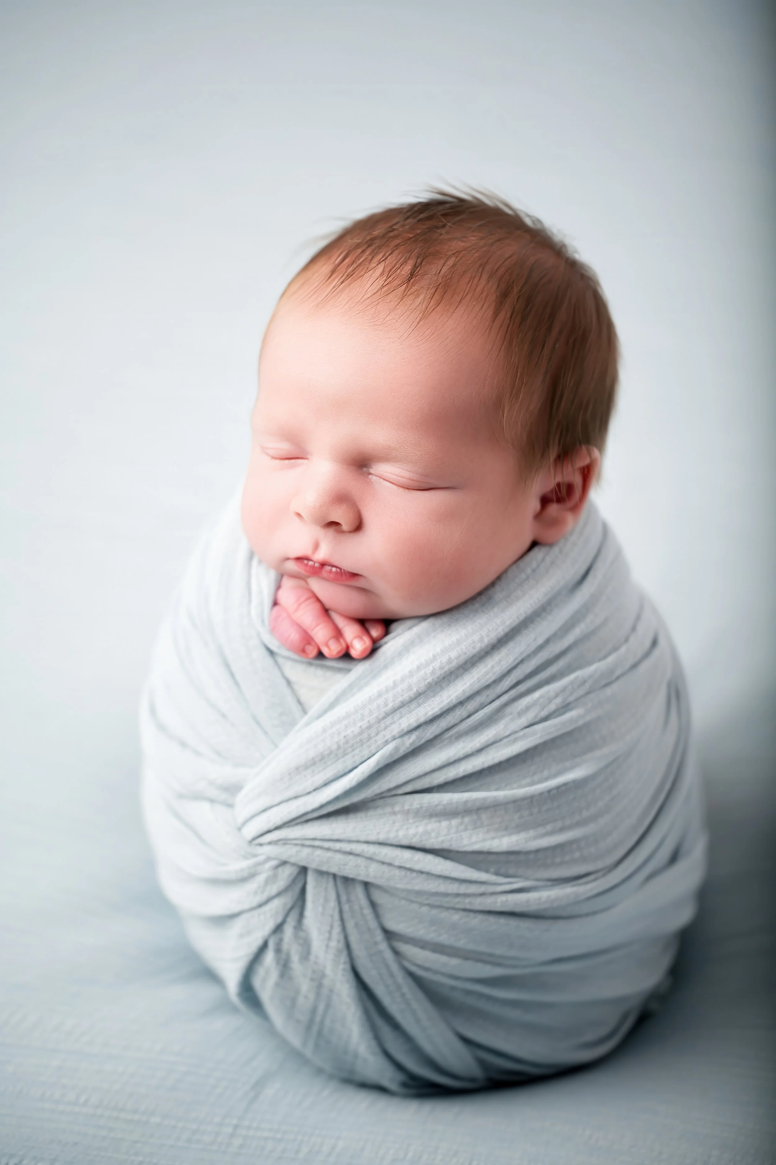 Close-up of a sleeping baby with fair skin and light brown hair, wrapped in a soft light gray blanket.
