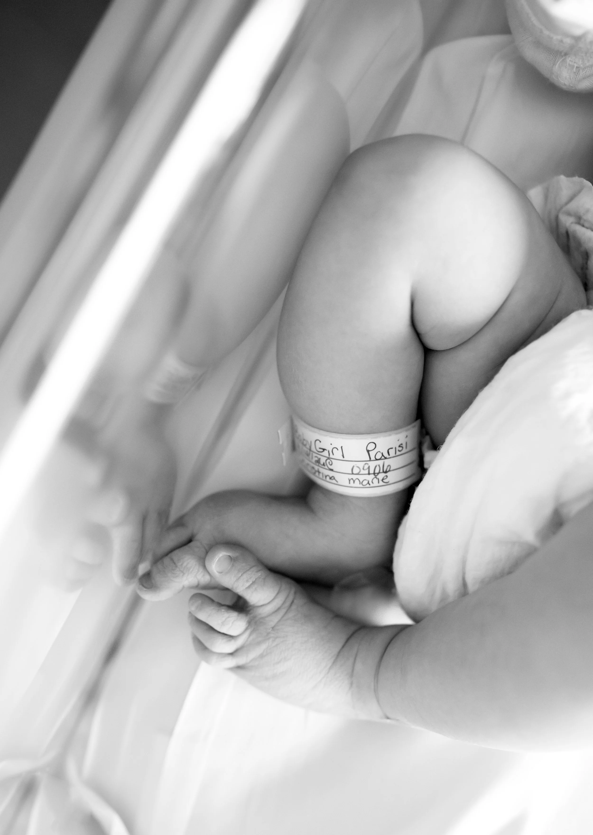 A newborn baby lying on a bed, with part of a person holding the baby. The baby is wearing a paper band with writing on it, and has a small arm raised. The image is in black and white.