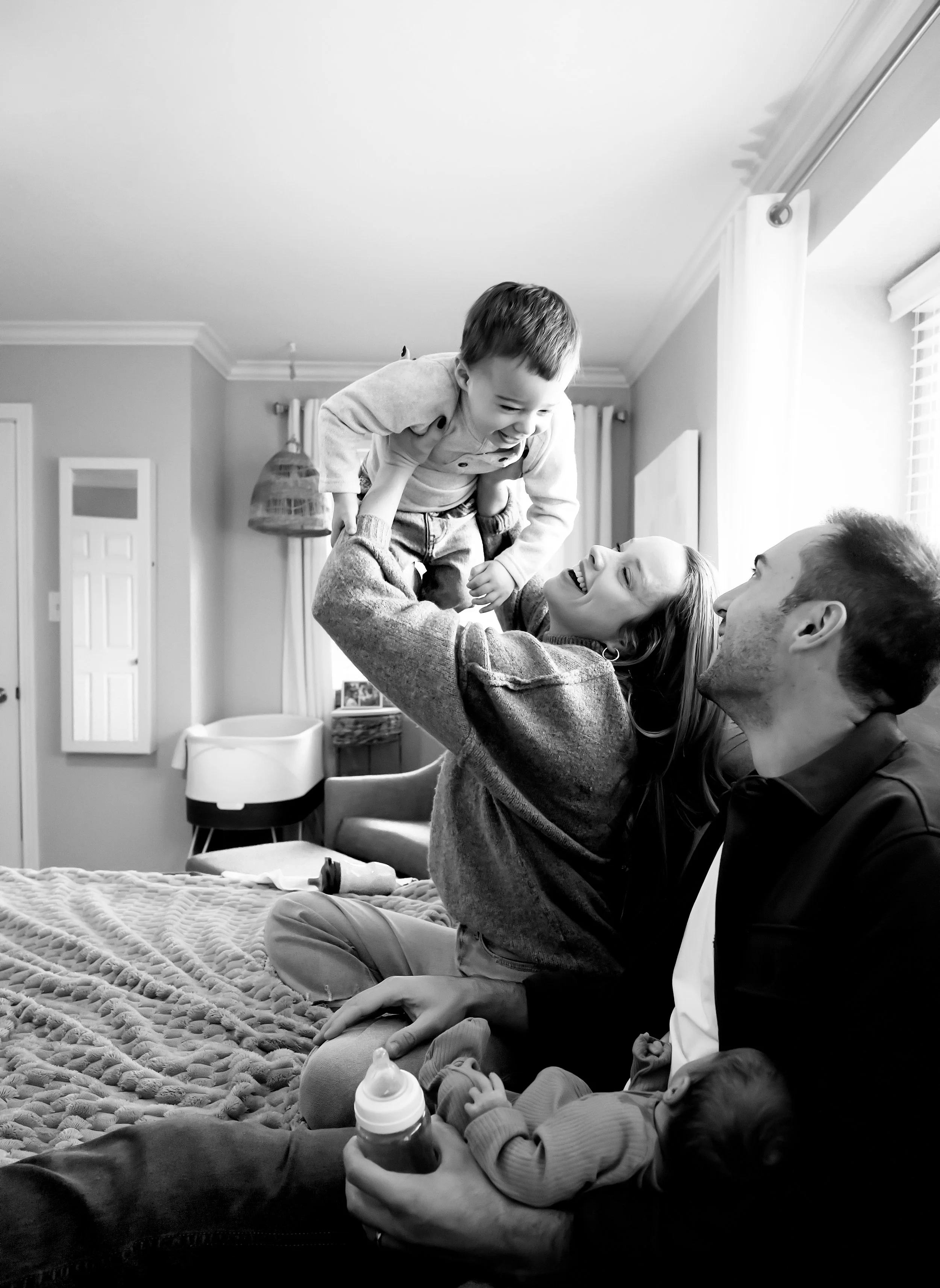 Family playing and smiling together in a cozy living room, with a woman lifting a young child into the air while another baby lies on a blanket, and a man and woman watching happily.