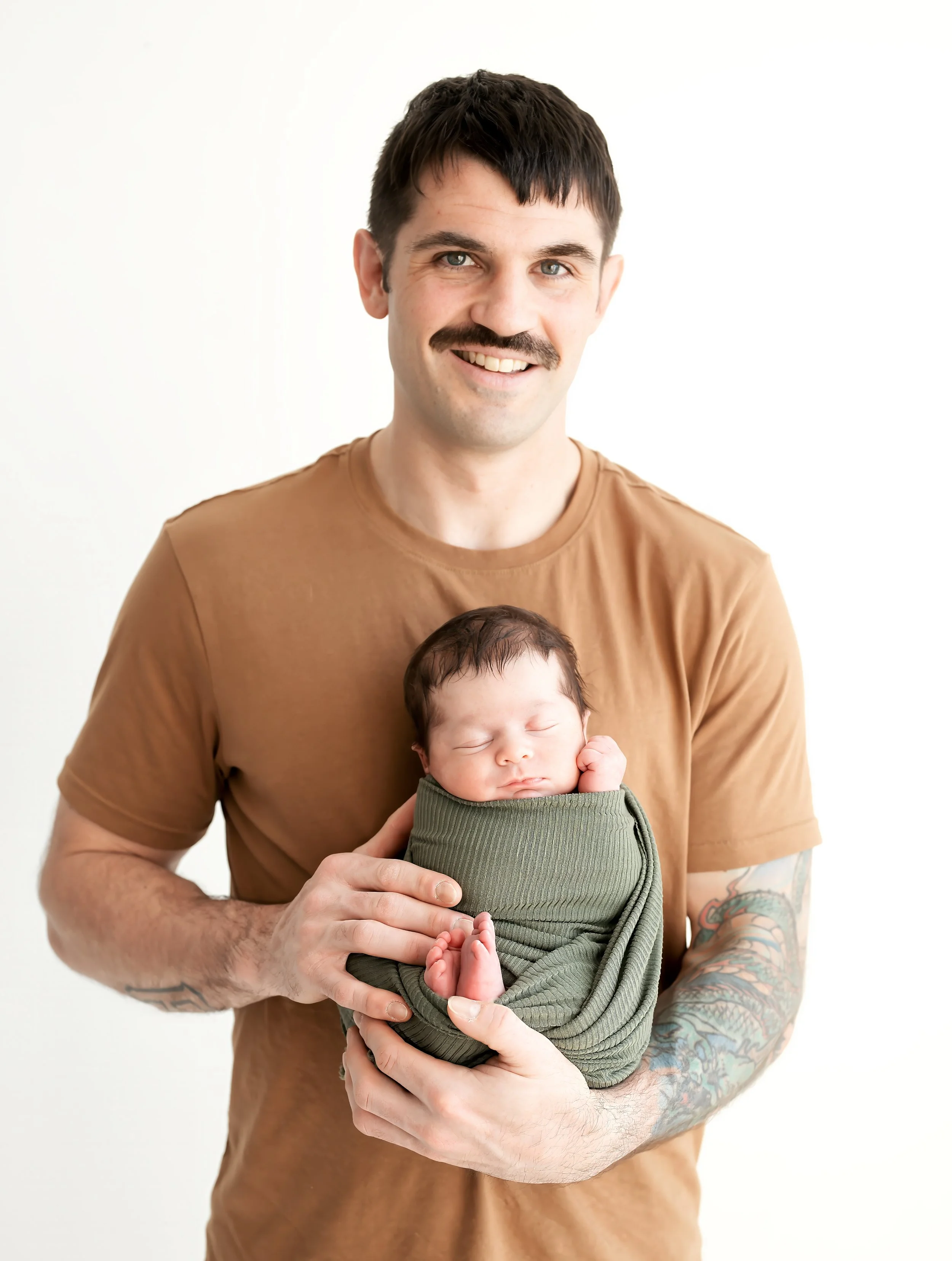 A man with a mustache and tattoos on his arms is smiling and holding a sleeping baby wrapped in a green blanket, against a white background.