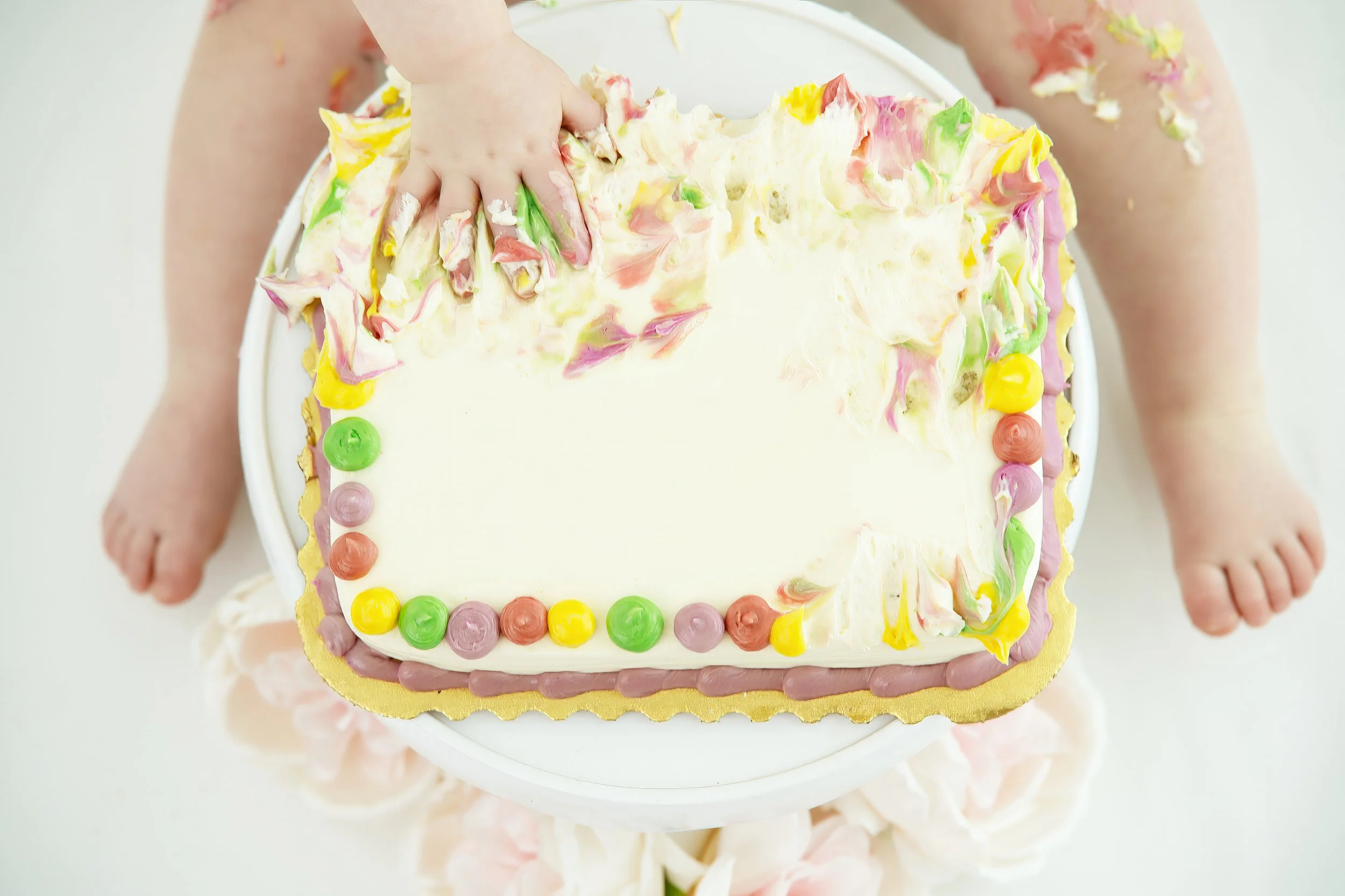 A child holding a rectangular birthday cake with white frosting, decorated with colorful icing blobs and a yellow border. The child's hands and arms are visible, with some frosting on their skin, and part of the child's legs and feet can be seen in t