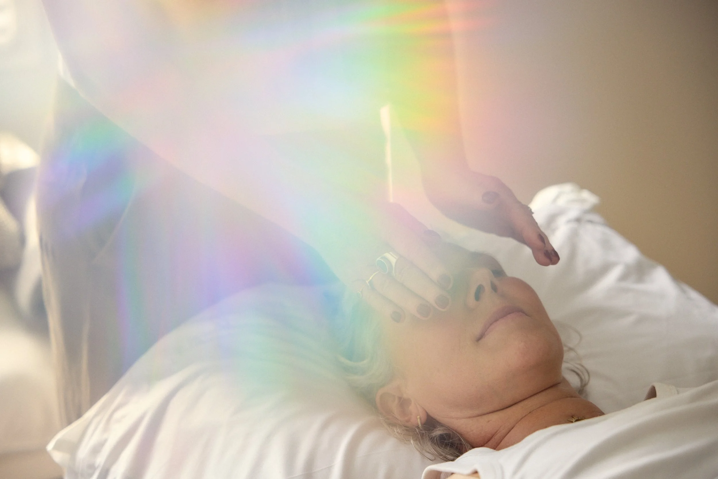 A woman with curly hair and a gold ring on her finger is lying on a bed, resting her head on a white pillow, with her eyes closed. The photo has a rainbow light effect across it.