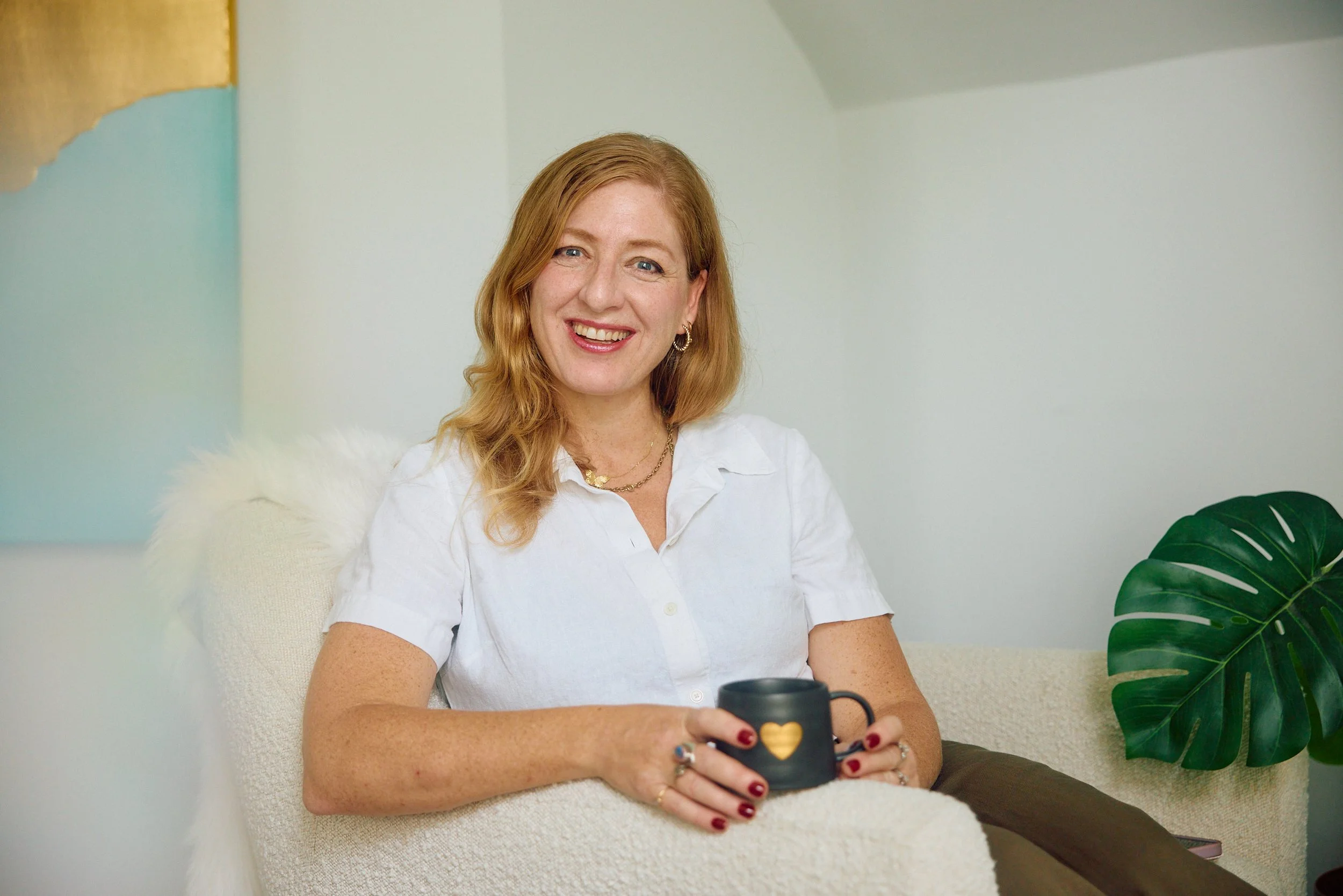 Woman with red hair, smiling, wearing a white shirt, holding a black mug with a gold heart, sitting in a cozy space with a large green plant and soft textured white chair.