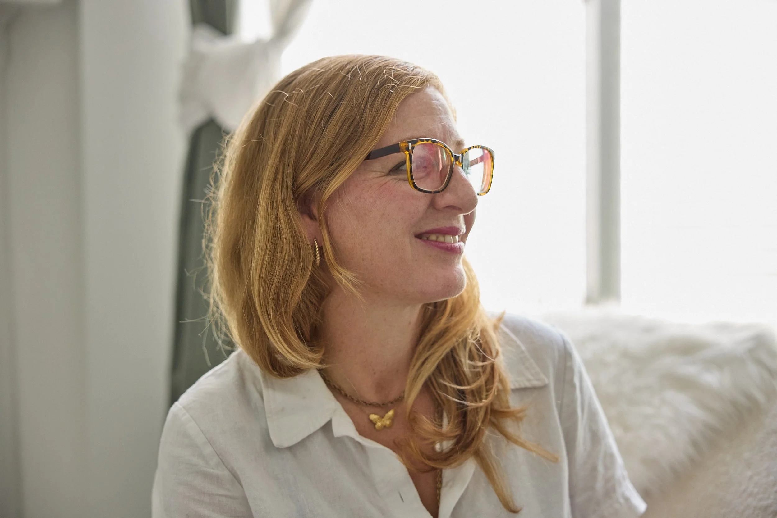 A woman with shoulder-length red hair and glasses with a tortoiseshell pattern, smiling, in a bright indoor setting.