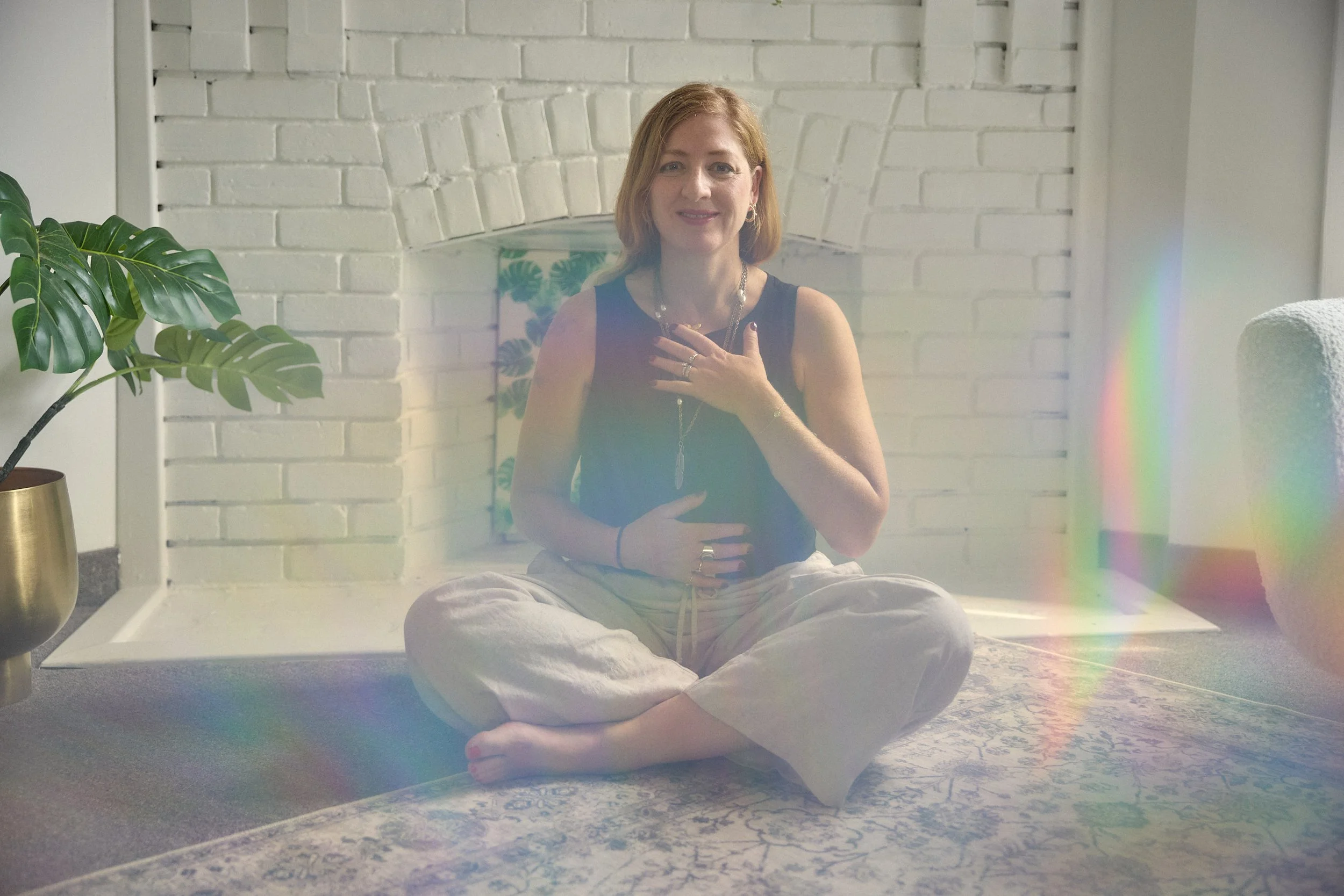 A woman with red hair sitting cross-legged on the floor in front of a white brick wall, smiling, with her right hand on her chest and her left hand on her stomach. There is a large green plant in a gold pot to her left and a colorful painting behind her. Sunlight creates a rainbow-like effect on the right side of the image.