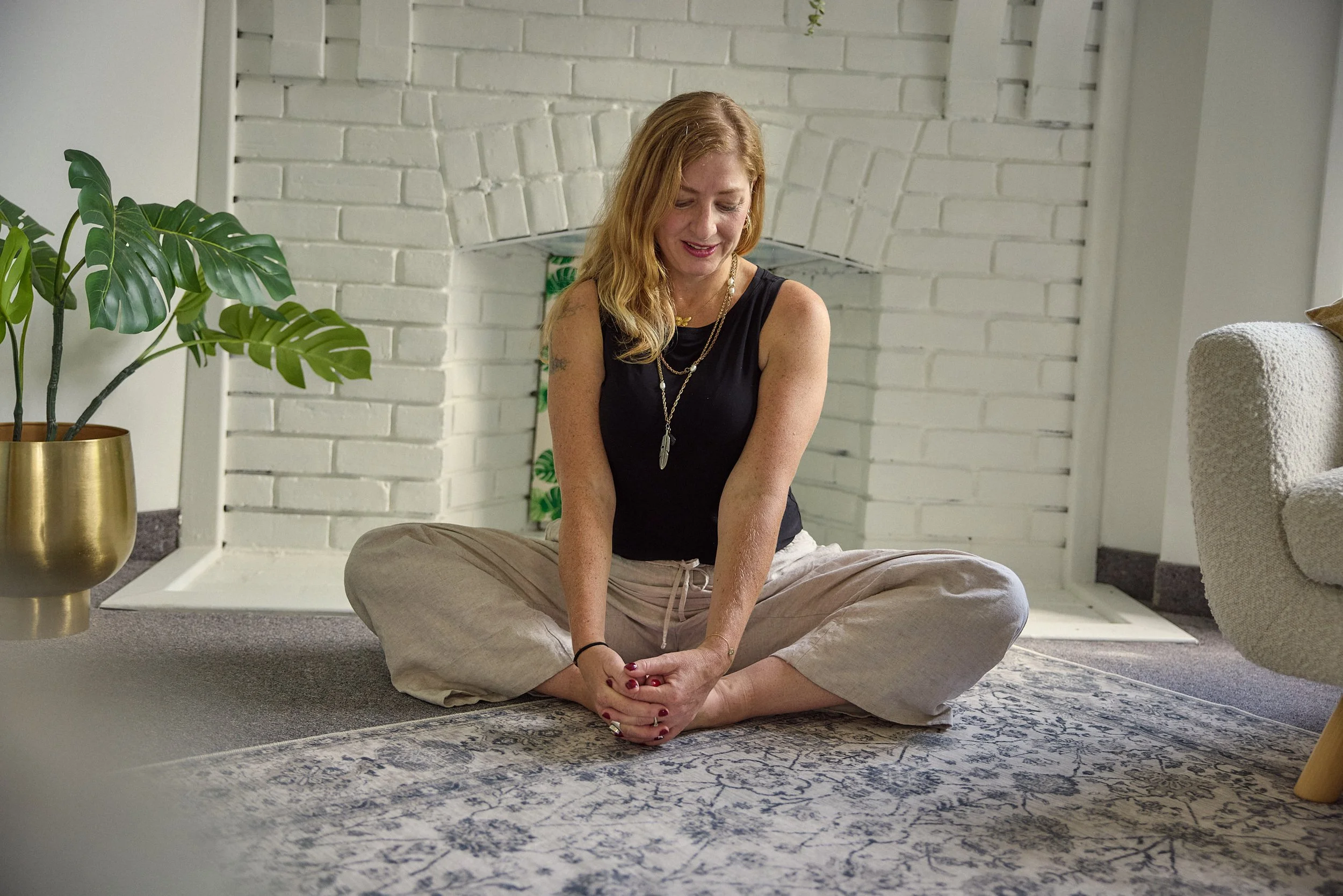 A woman with red hair sitting on a patterned rug in a room with white brick walls, a potted plant in a gold vase nearby, and a beige armchair. She is wearing a black sleeveless top and beige pants, smiling and looking down.