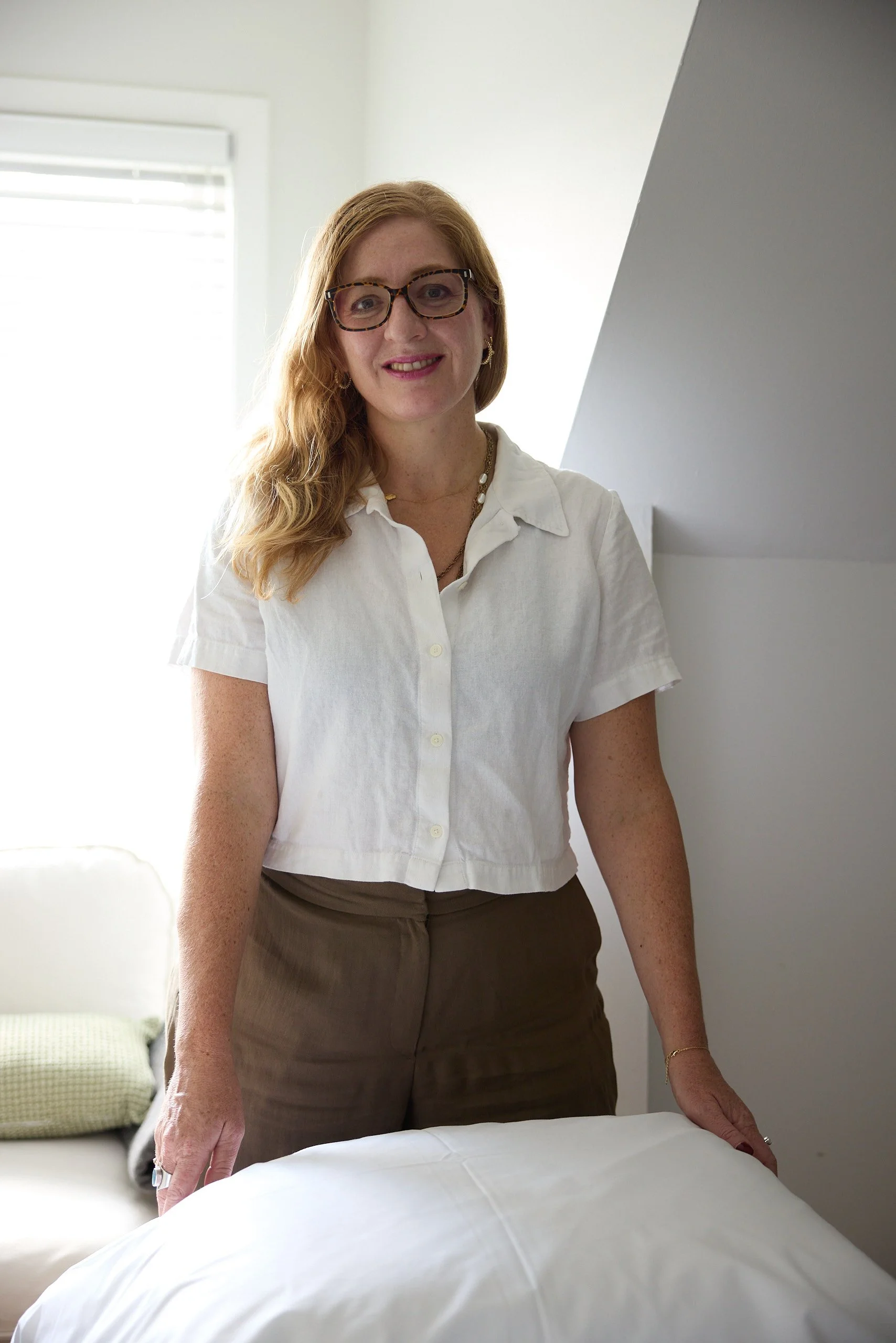 A woman with long red hair, glasses, and jewelry is standing beside a bed, smiling. She is wearing a white shirt and brown pants, with her hand resting on the bed.