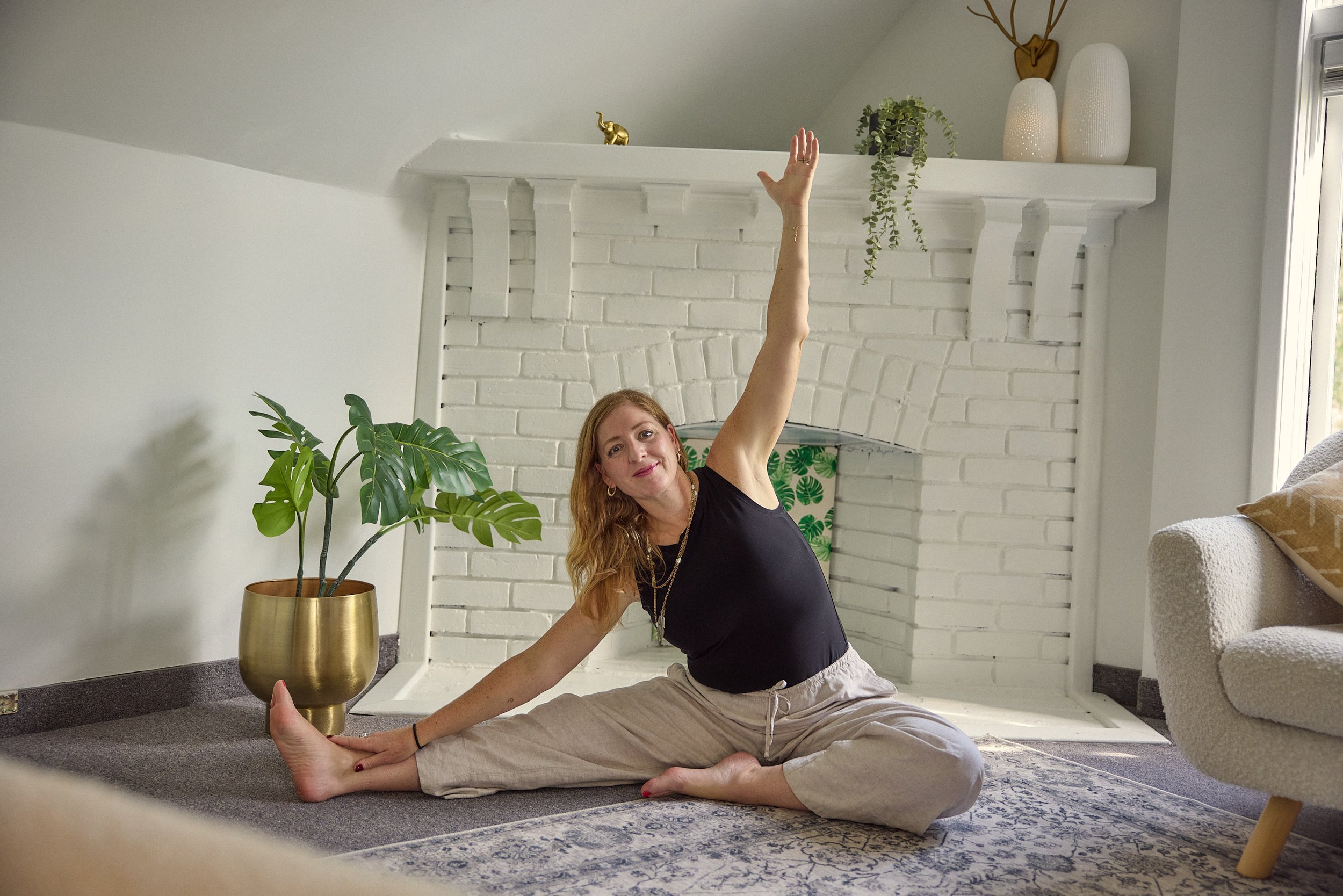 A woman with red hair practicing yoga indoors, seated on the floor in a split pose, with one arm raised and the other arm resting on her foot, near a large potted plant and a white brick fireplace.