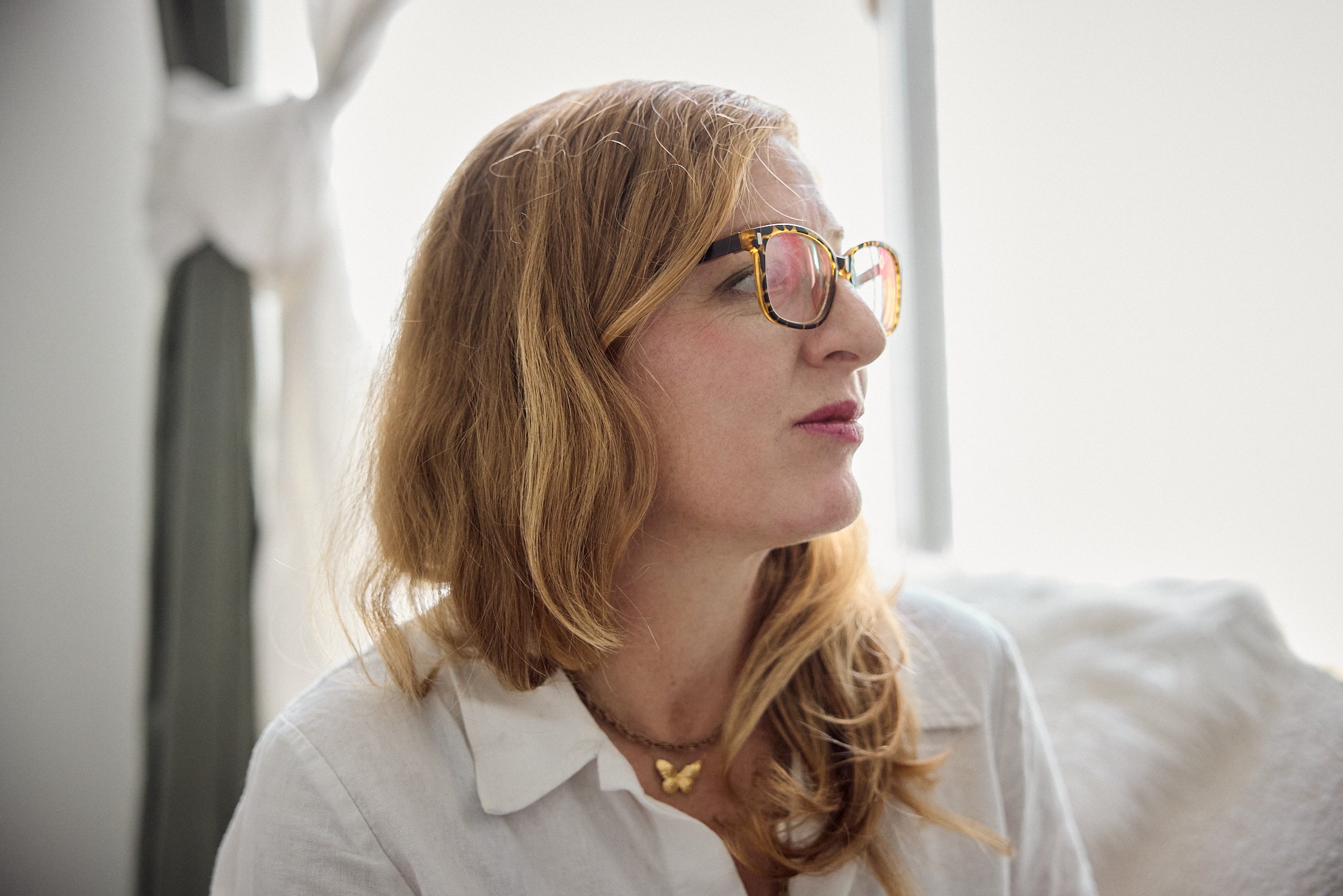 A woman with red hair wearing tortoiseshell glasses and a white shirt, looking to the side.