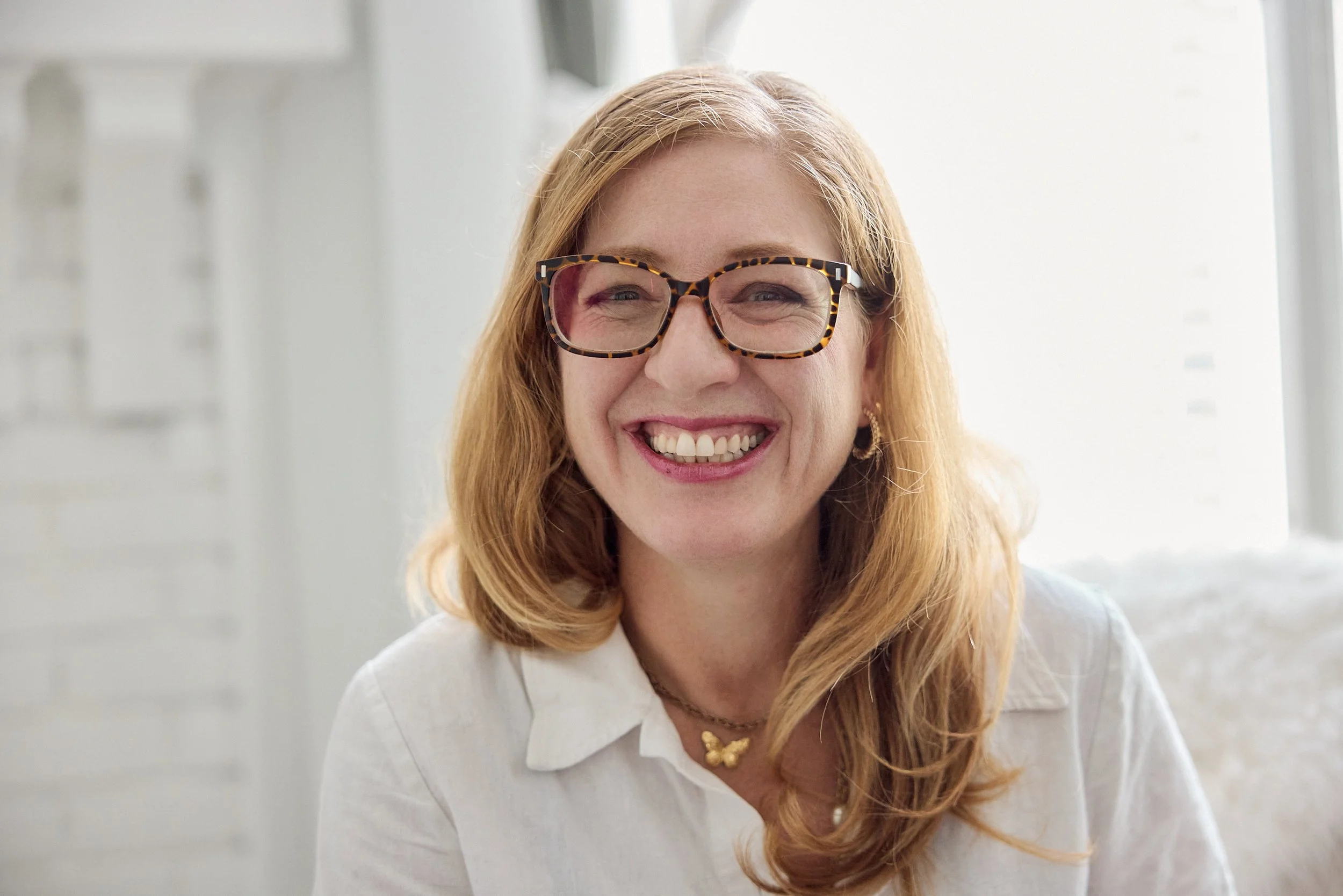 Close-up of a woman with red hair, wearing tortoiseshell glasses, smiling and showing her teeth, in a bright room with white walls.