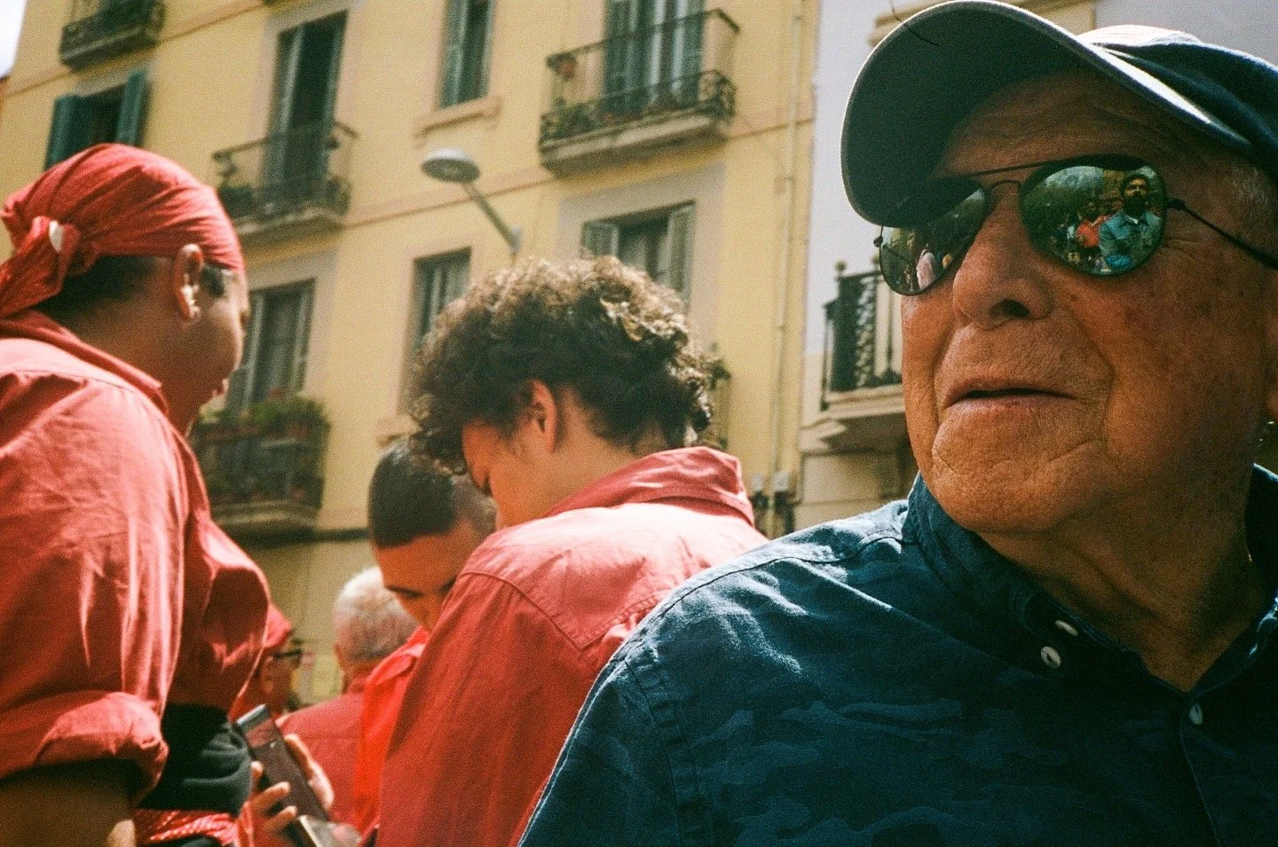 Close-up of an elderly man wearing sunglasses and a cap, with a group of people in the background, outdoors in front of a yellow building with balconies.