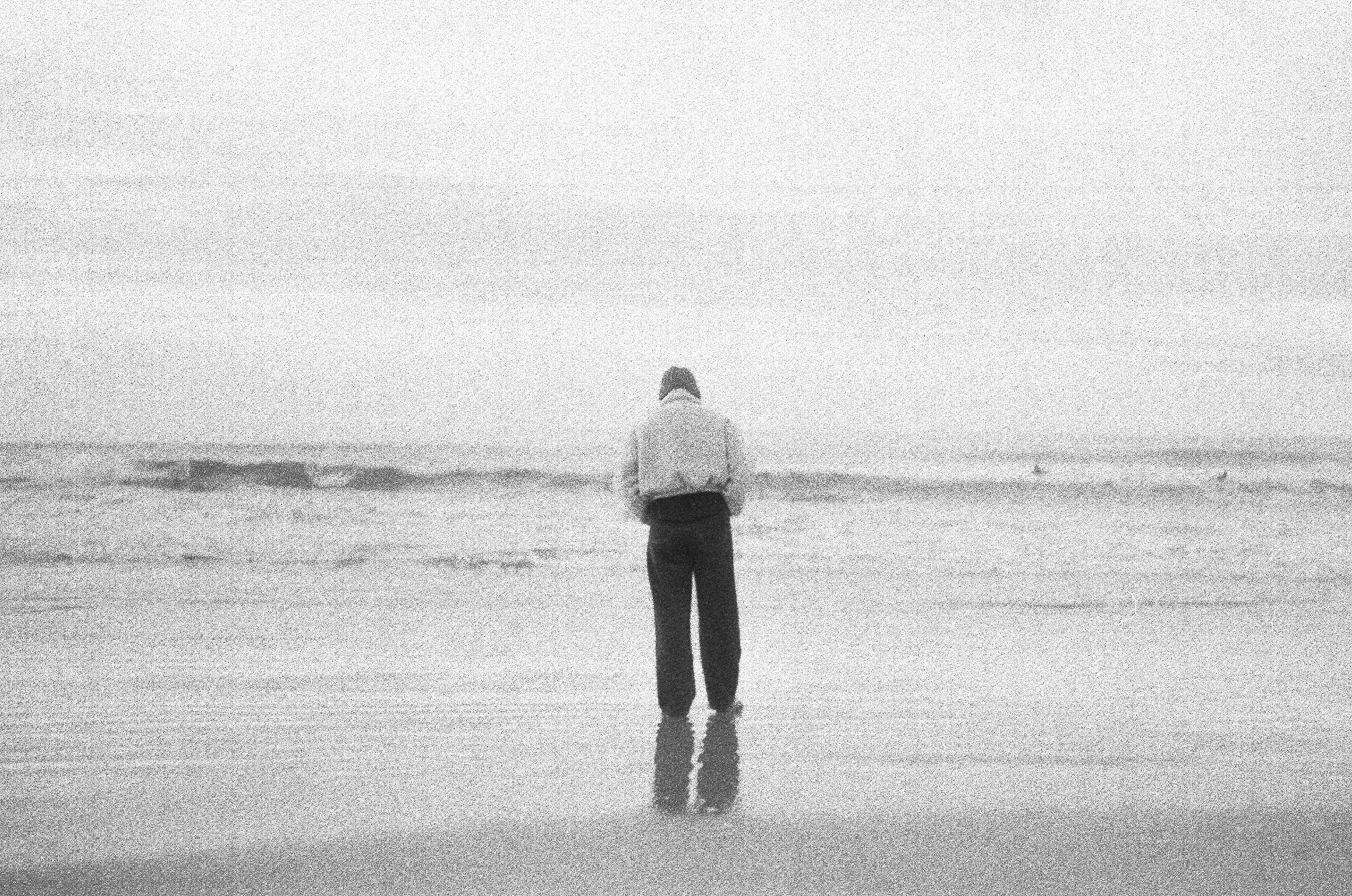 A person standing alone on a beach facing the ocean, with their back to the camera and their reflection visible on the wet sand.