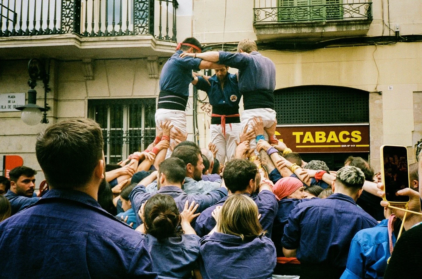 Crowd of people participating in a traditional human tower event, with two individuals standing on the shoulders of others, forming a multi-layered human tower as part of a cultural festival.