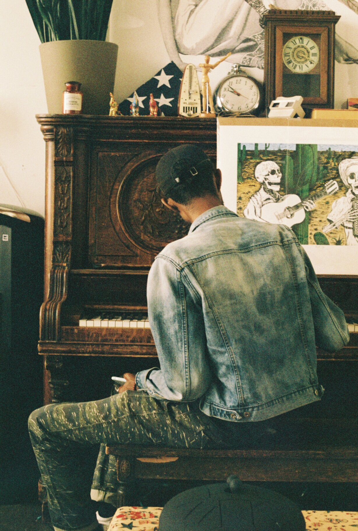A person wearing a black cap, denim jacket, and patterned pants, sitting on a wooden bench and looking at their phone. They are in front of an antique wooden piano with a decorative carving on its front panel. Behind them, there are artwork, a painti