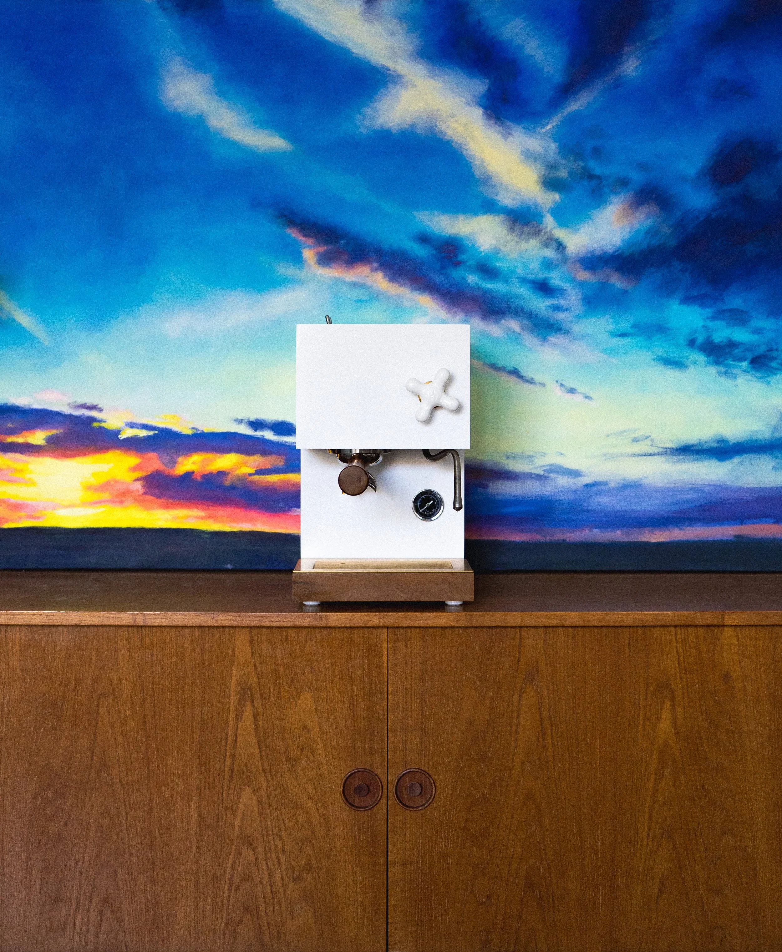 A white machine with a small white cross-shaped object on top, a gauge, and other mechanical parts, positioned in front of a colorful sunset sky mural with clouds.