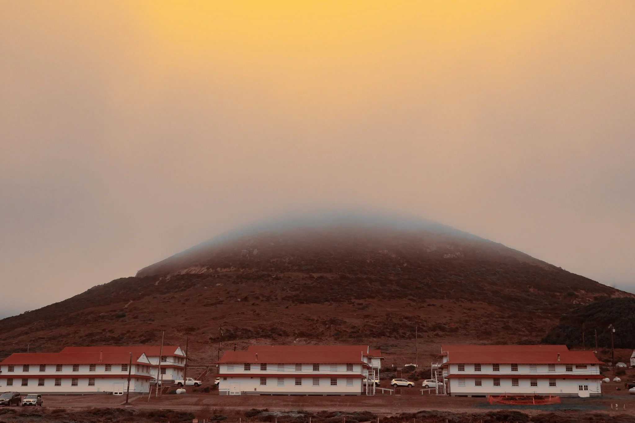 A mountain in the background with mist at its peak, above reddish-brown terrain, and a row of white buildings with red roofs at the base, along with parked cars and utility poles.