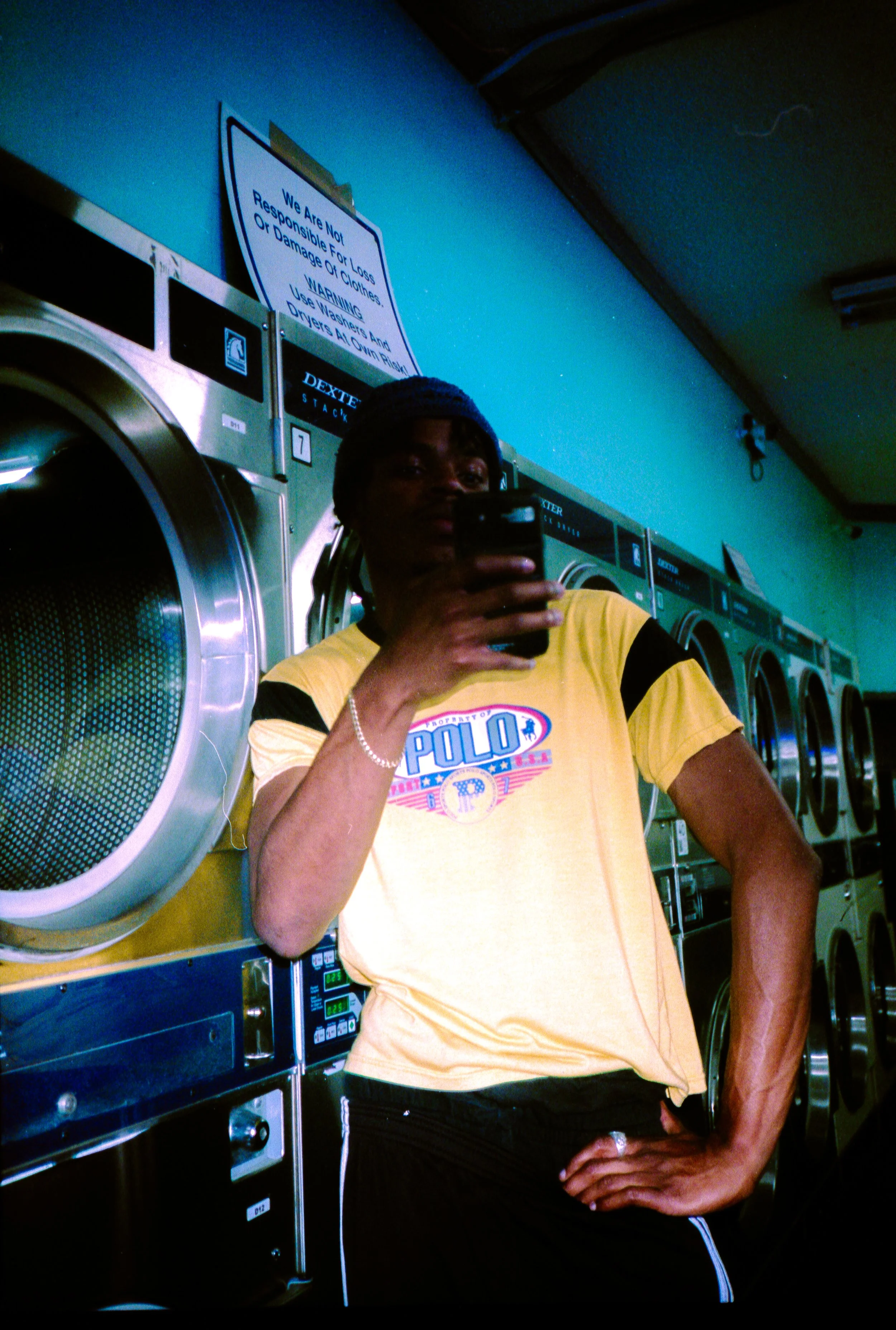 A person taking a mirror selfie in a laundromat, standing in front of a row of washing machines, wearing a yellow t-shirt with a Polo logo, a beaded bracelet, and a dark hat.