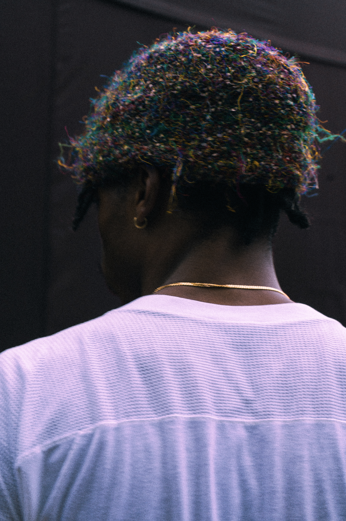 Back of a person with multicolored, textured hair wearing a white shirt and gold jewelry.
