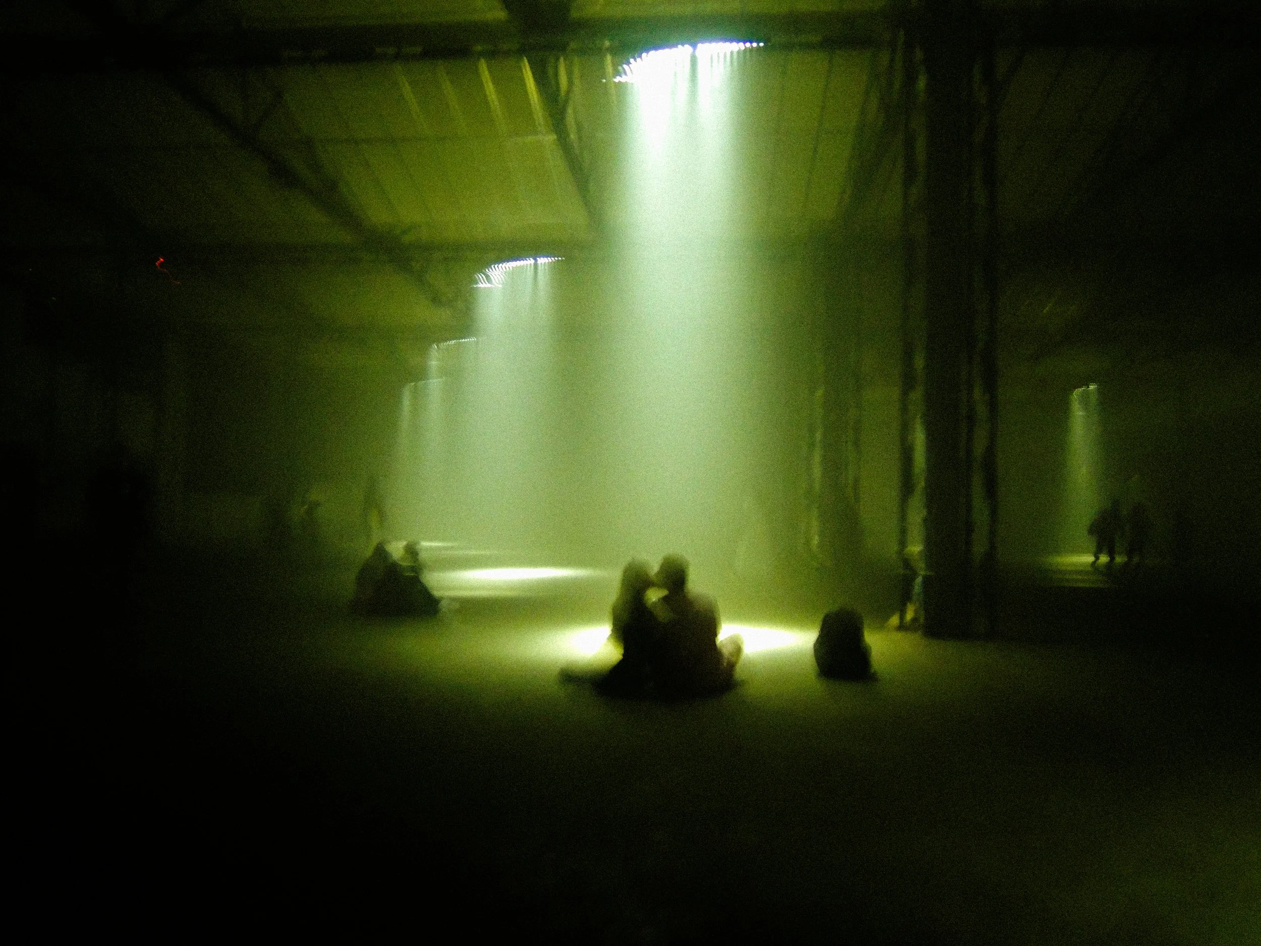 People sitting on the ground in front of a stage with a green light shining from above, in a dark room or arena.