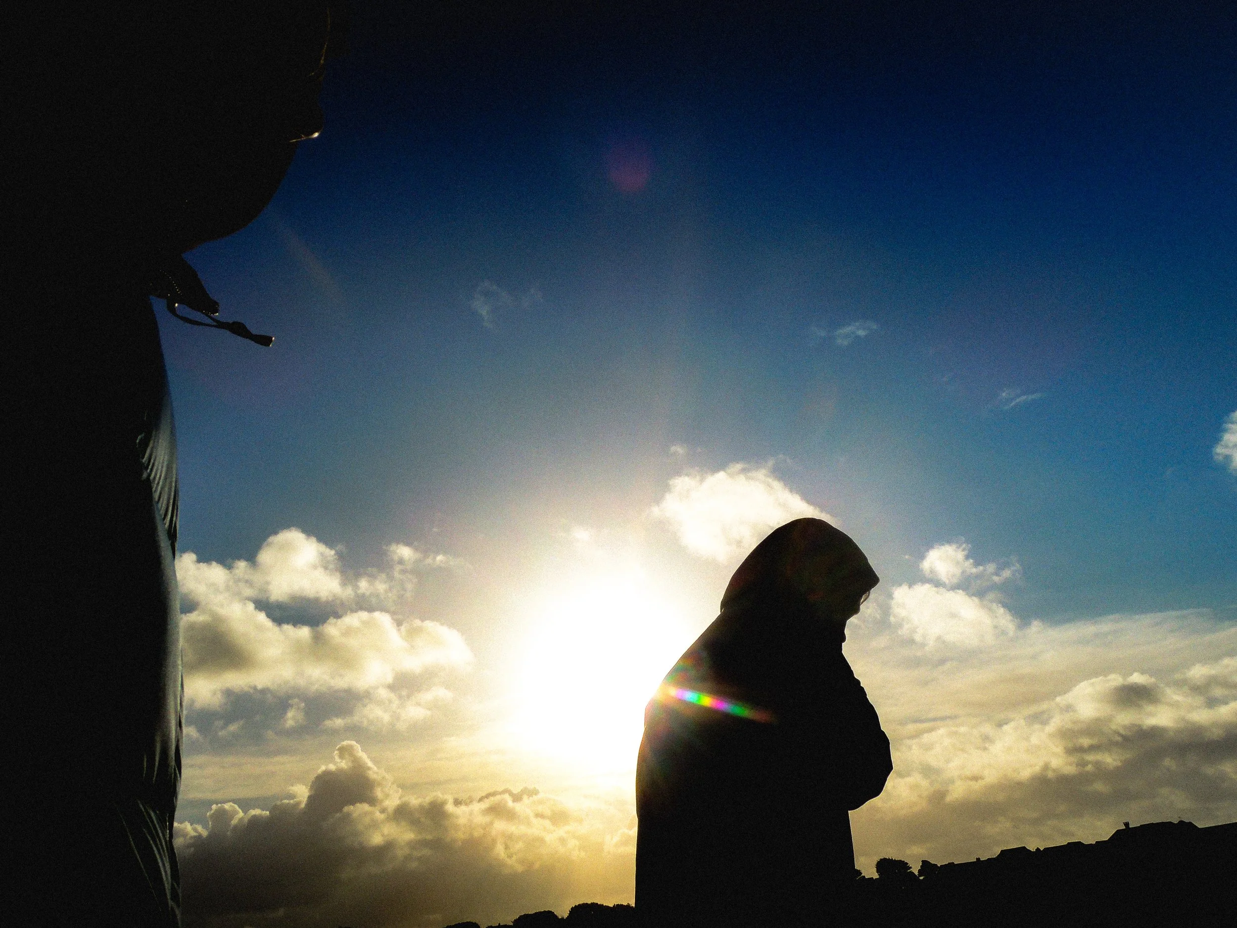 Silhouettes of two people, one on the left and one on the right, against a bright sky with clouds and the sun near the horizon.