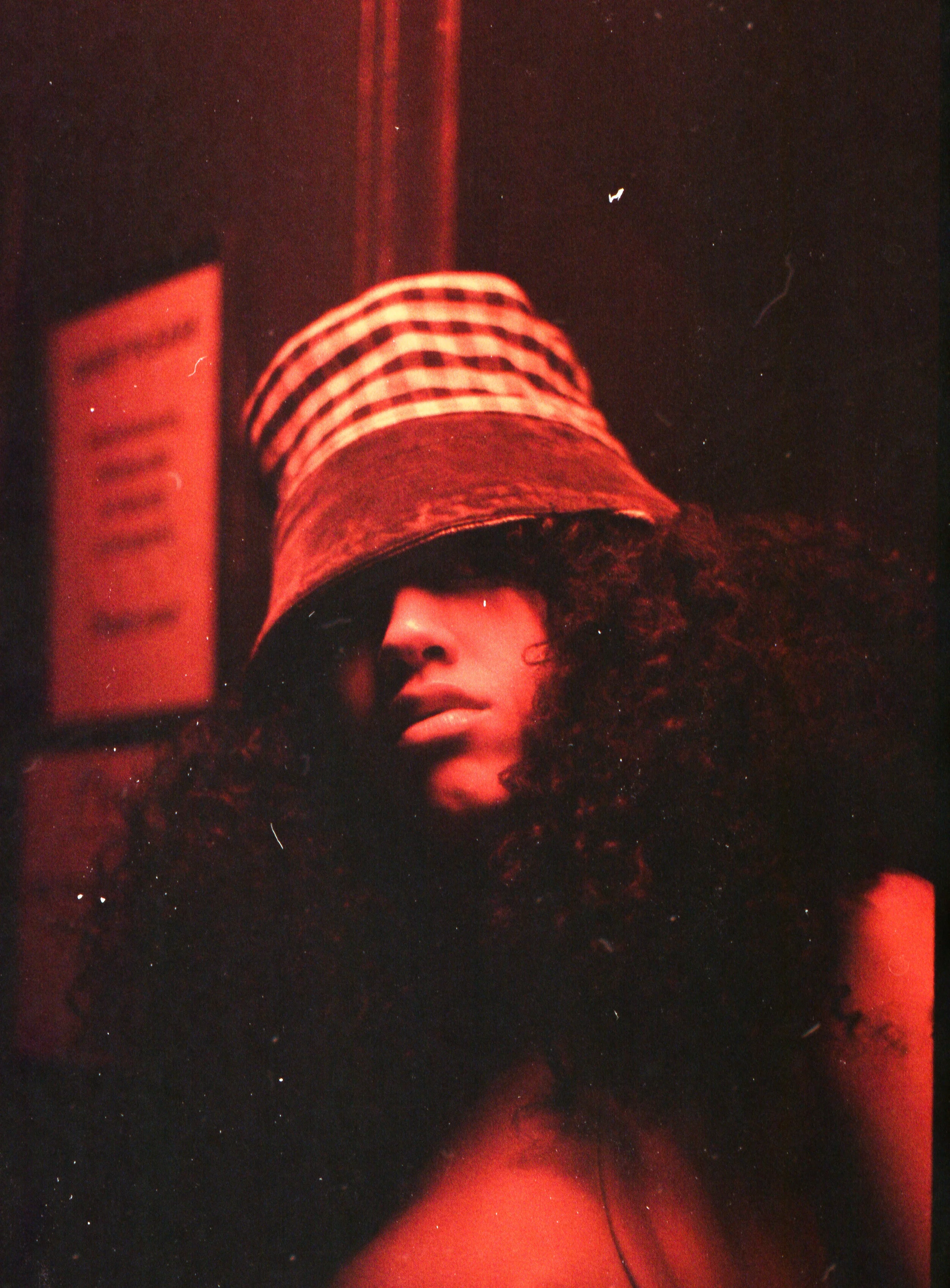 Person with curly hair wearing a striped hat, face partially visible, photo taken with red lighting in a dark environment.