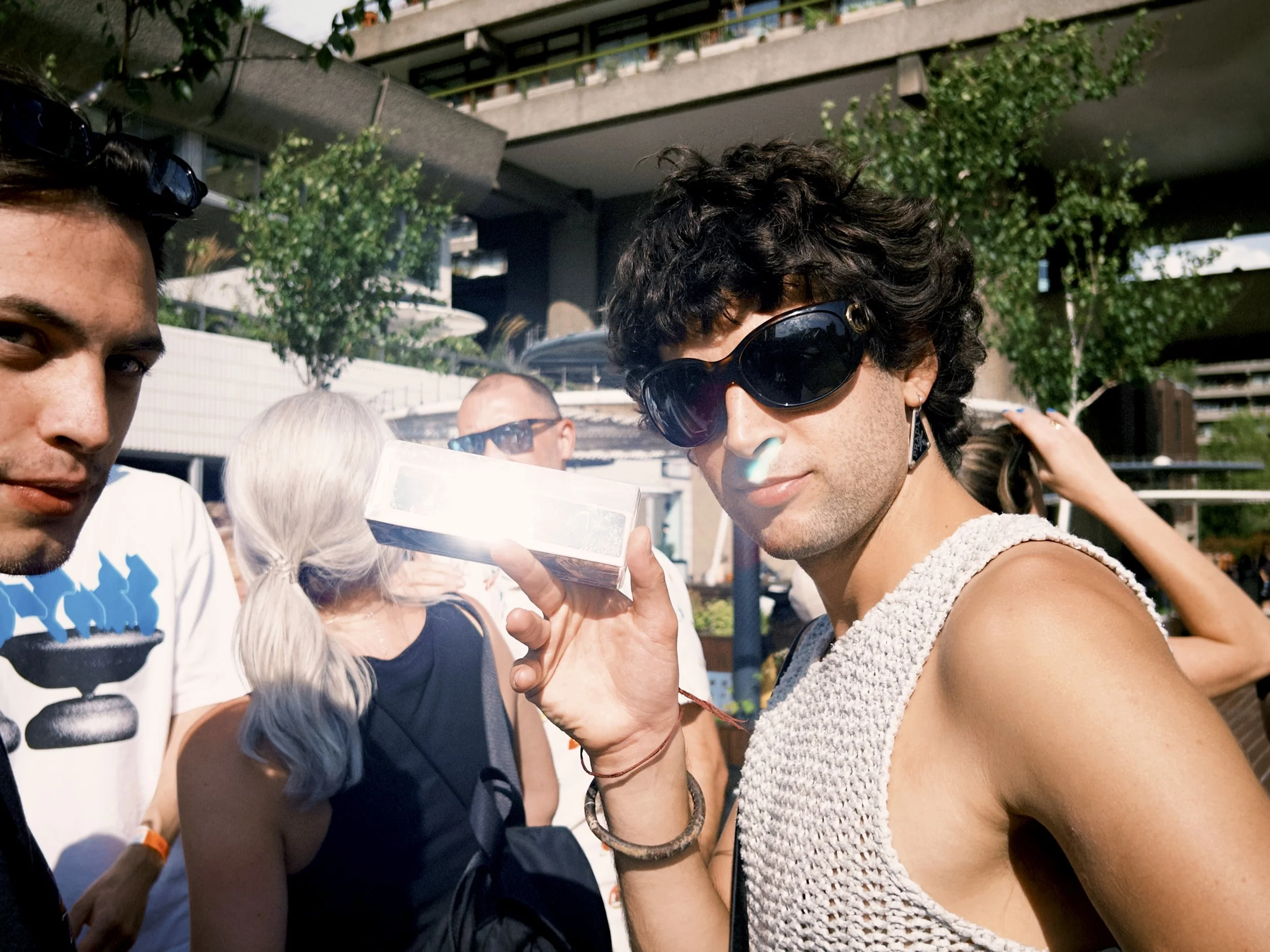Two young men are standing outdoors in sunlight; one has curly dark hair, wearing large sunglasses and a sleeveless top, while the other has dark hair and is wearing a white shirt with a graphic design.